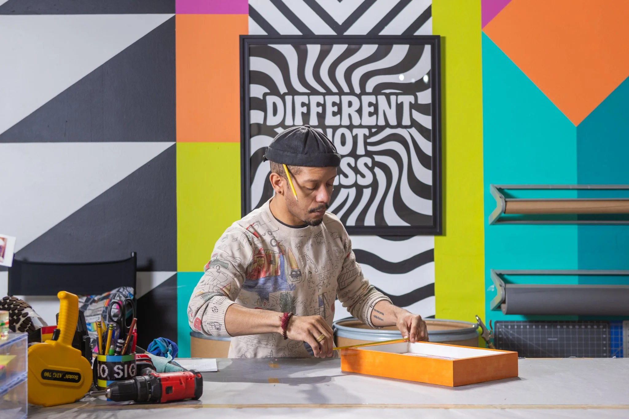 A person with short hair and glasses working on a project at a cluttered worktable, with tools and supplies in the foreground, and colorful geometric wall art including a black and white striped background with the words "Different Not Less" behind.
