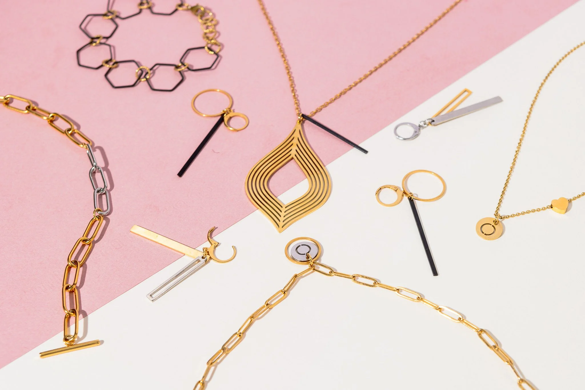 Assorted gold and silver jewelry including chains, rings, and earrings arranged on a pink and white background.