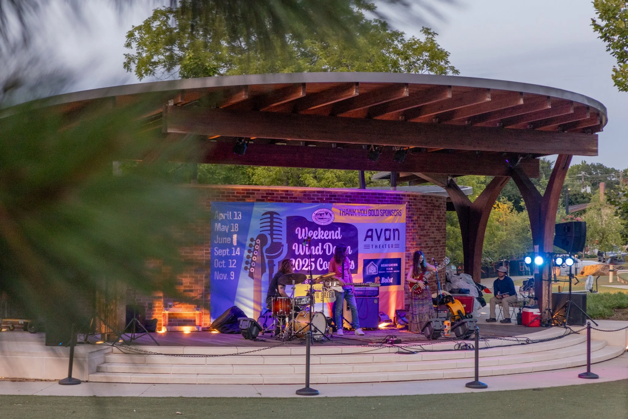 An outdoor stage with a band performing at night, with a banner promoting a Wind Day event and a brick building in the background. Trees frame the scene, with spotlights illuminating the stage.