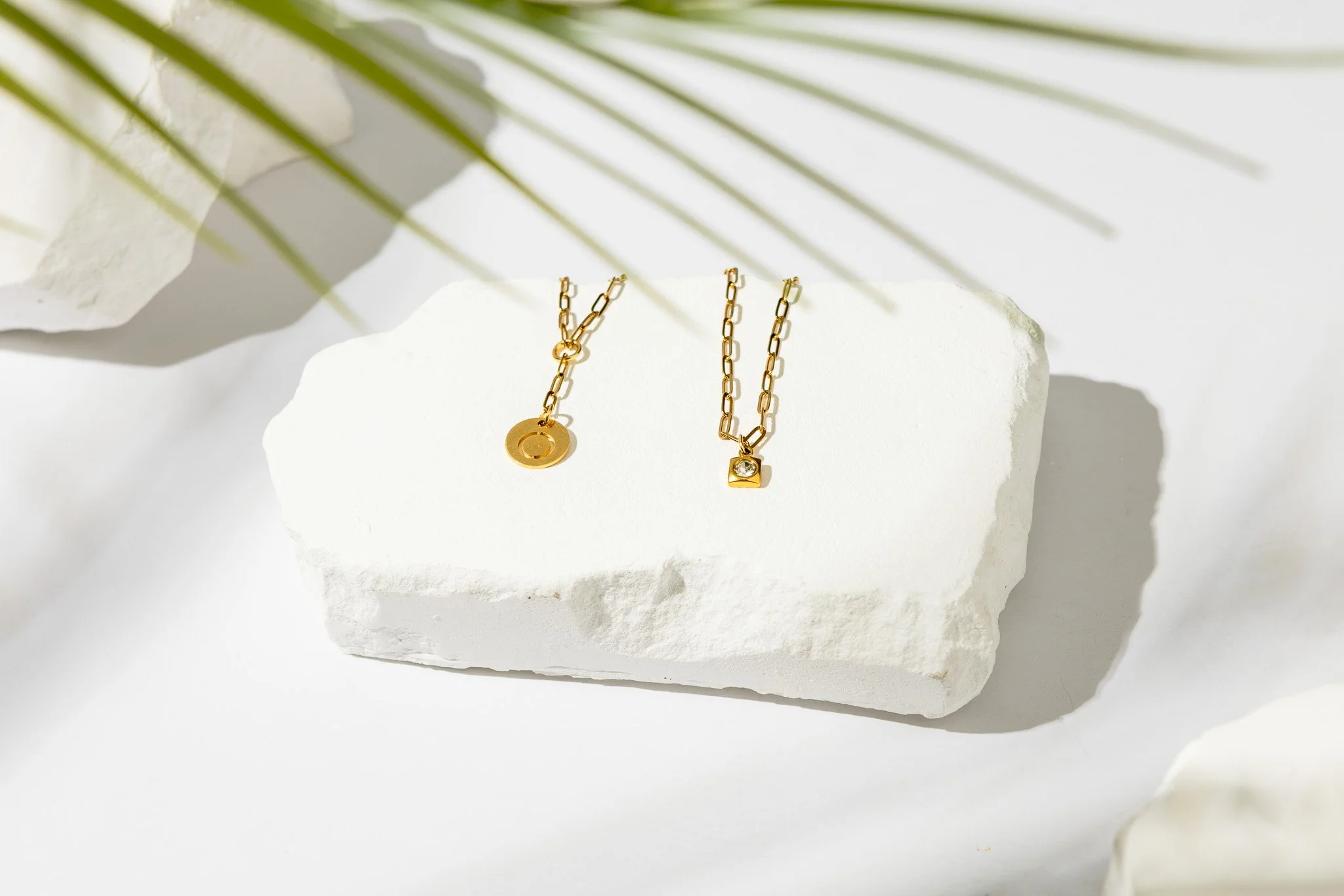 A pair of gold chain necklaces with pendants displayed on a white textured surface, with shadows and palm leaves partially covering the scene.