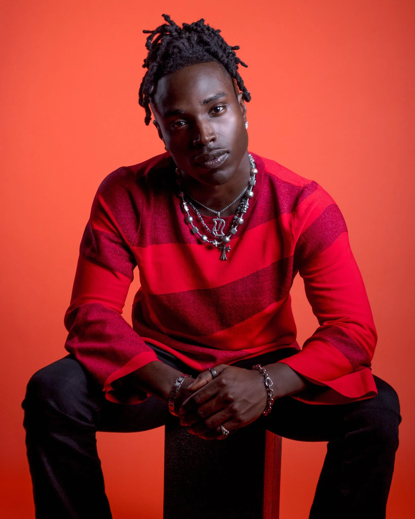 A young man with dark skin and styled dreadlocks poses against an orange background. He is wearing layered necklaces, rings, and earrings, and a red and black striped sweater. He looks directly at the camera with a neutral expression.