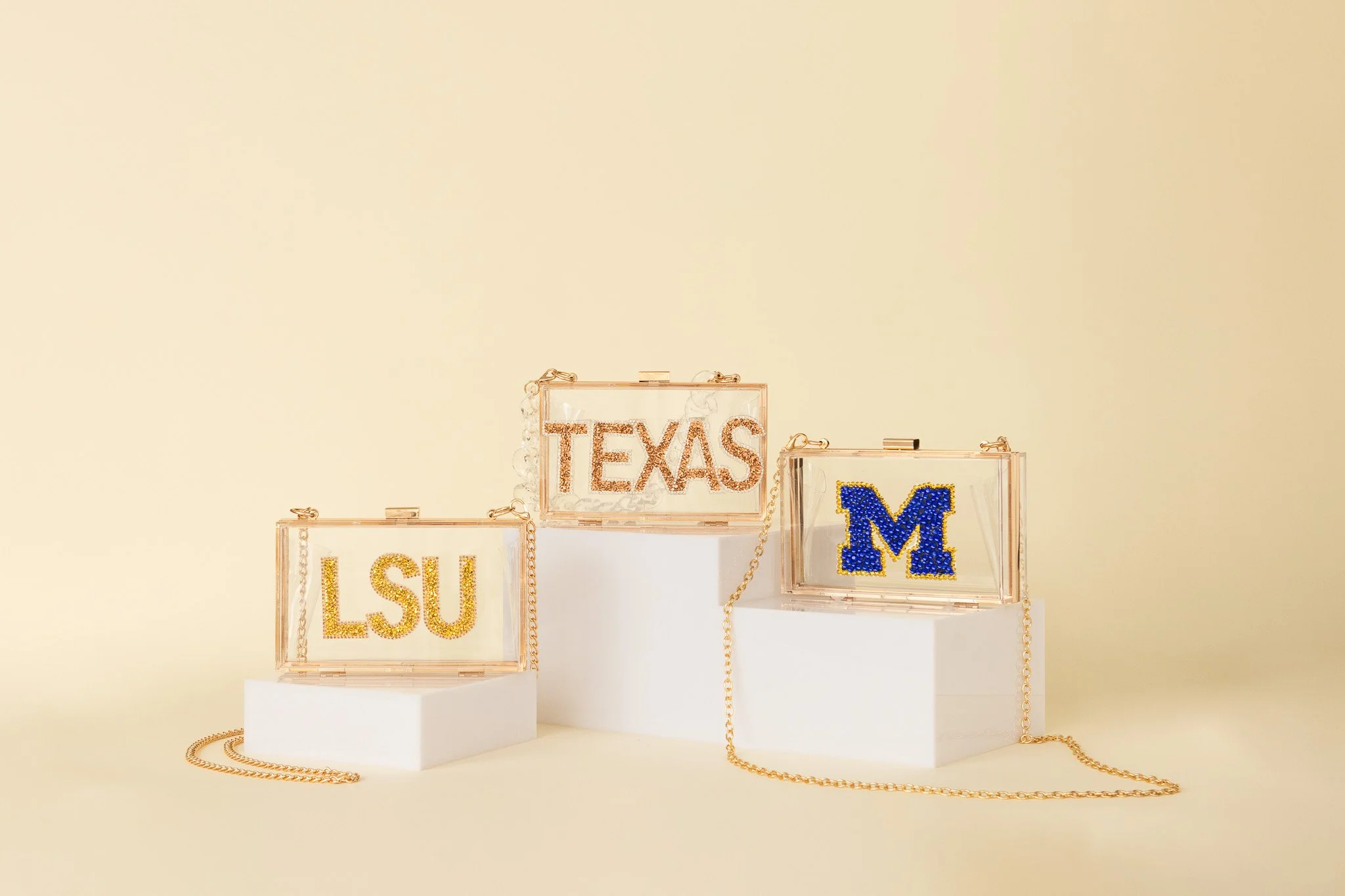 Three transparent acrylic boxes displaying university logos: LSU in yellow rhinestones, Texas in pink rhinestones, and Michigan in blue rhinestones, with gold chains hanging from some boxes, on white pedestals against a plain beige background.
