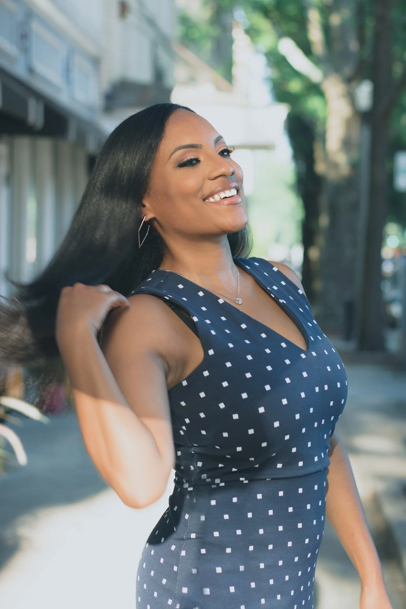 A woman with long black hair smiling outdoors in a sleeveless navy blue polka dot dress, with trees and buildings in the background.