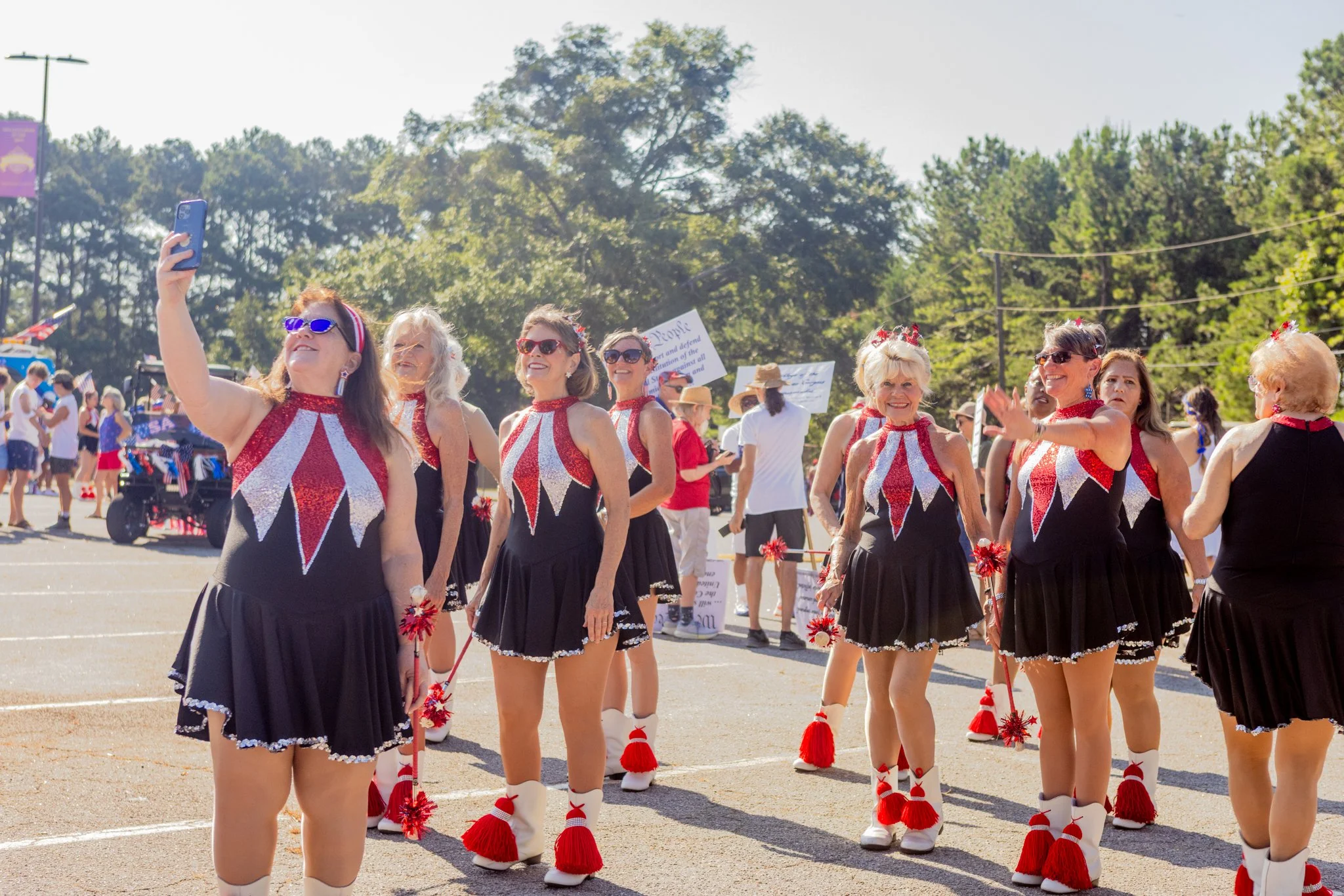 A group of women dressed in cheerleader uniforms with red, white, and black colors, standing outdoors at a parade or celebration. Some women are wearing sunglasses, and one woman is taking a selfie. There are other people and signs in the background,