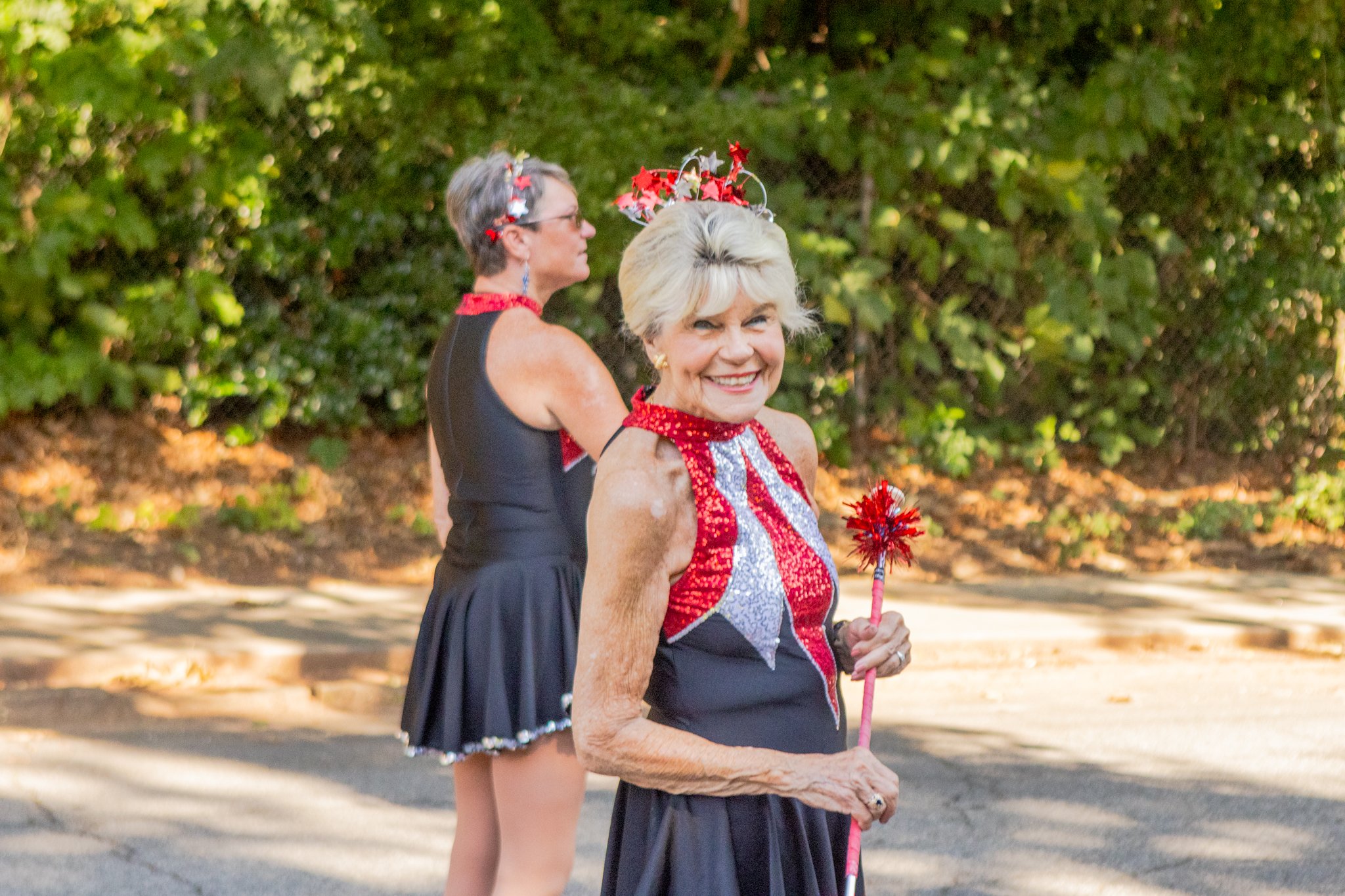 Two women, one elderly and one younger, dressed in patriotic red, white, and blue outfits, celebrating outdoors with greenery in the background. The elderly woman has a red, white, and blue headpiece and a matching top, and is smiling while holding a