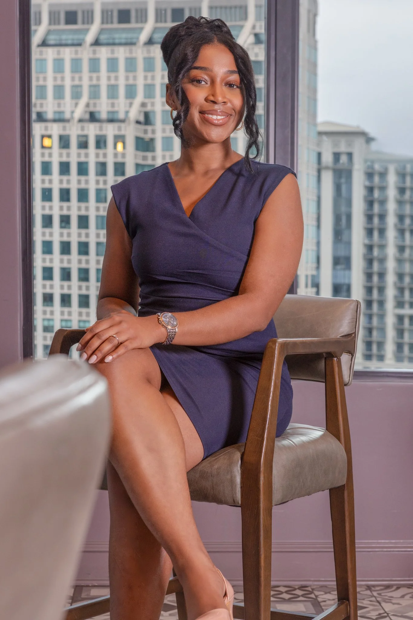 A woman in a navy blue dress sitting on a chair, smiling, with a city skyline visible through a window behind her.