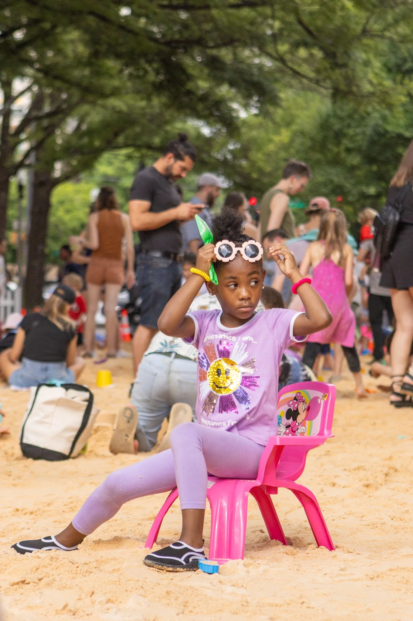 A young girl sitting on a small pink chair in a park on a sandy surface, adjusting her sunglasses with her hands. She is wearing a purple T-shirt with a colorful design, purple leggings, and black shoes. The background shows several people, some standing and others seated, with greenery and trees overhead.