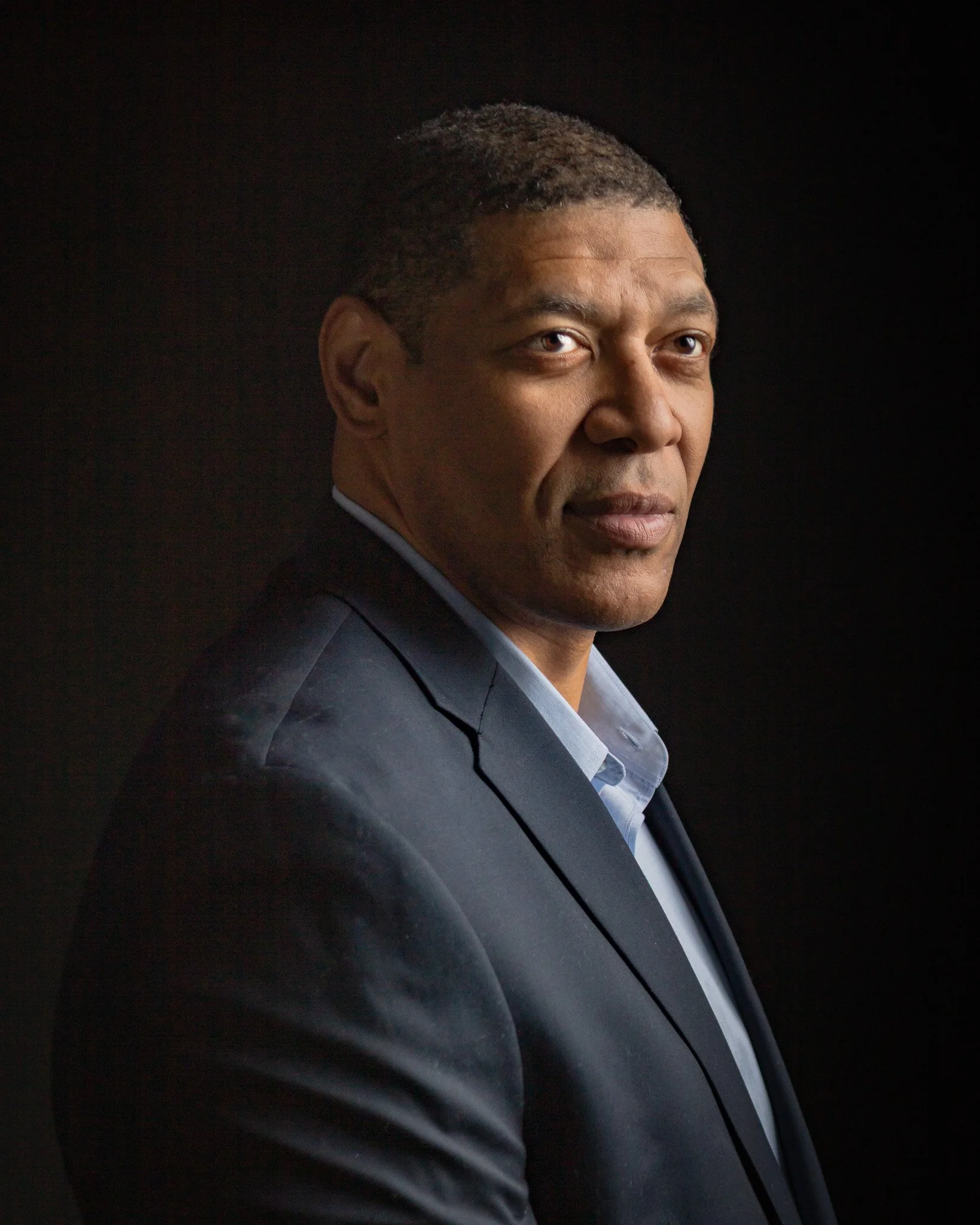 A man in a dark suit with a light blue shirt looking at the camera against a black background.