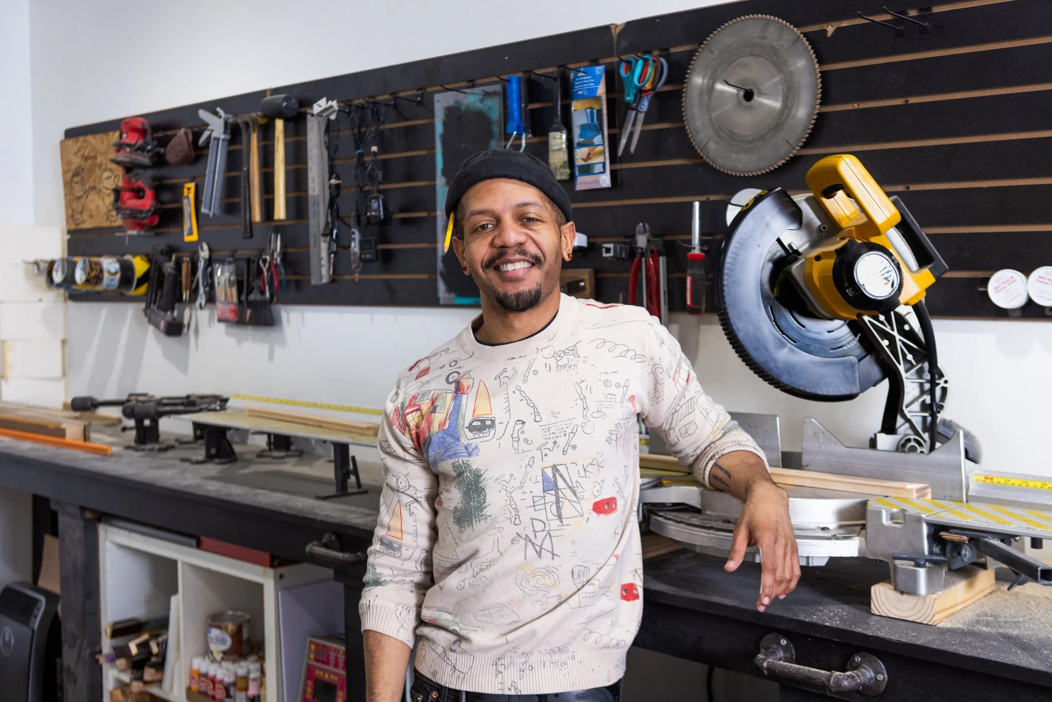 A man smiling in a workshop surrounded by tools hung on a board and a yellow miter saw.