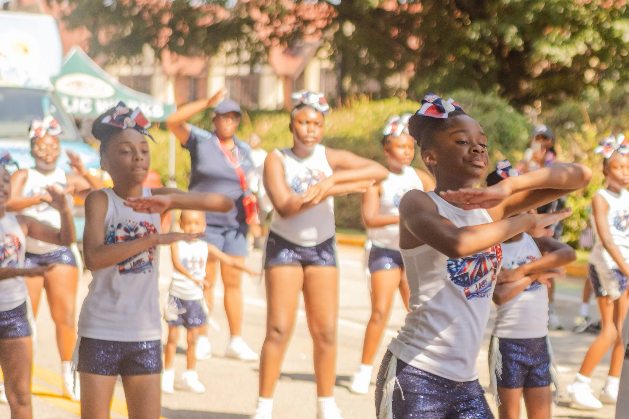 A group of young girls performing a dance routine outdoors, wearing matching white and blue costumes with patriotic-themed hair bows, during a daytime event.