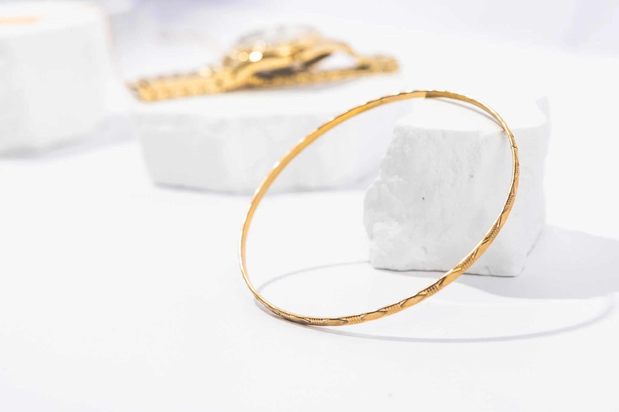 A single gold hoop earring placed on a white textured stand, with blurred gold jewelry in the background on a white surface.