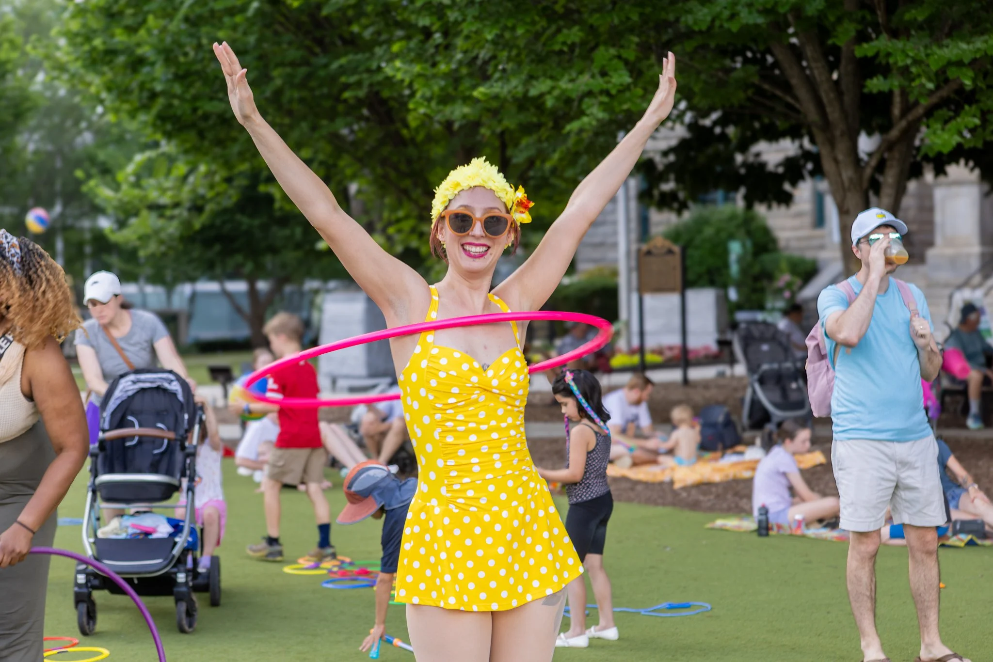 A woman wearing an orange swimsuit with yellow polka dots, a yellow flower headband, sunglasses, and pink lipstick is hula hooping at an outdoor event with children and other adults, some sitting on the grass and some walking around.