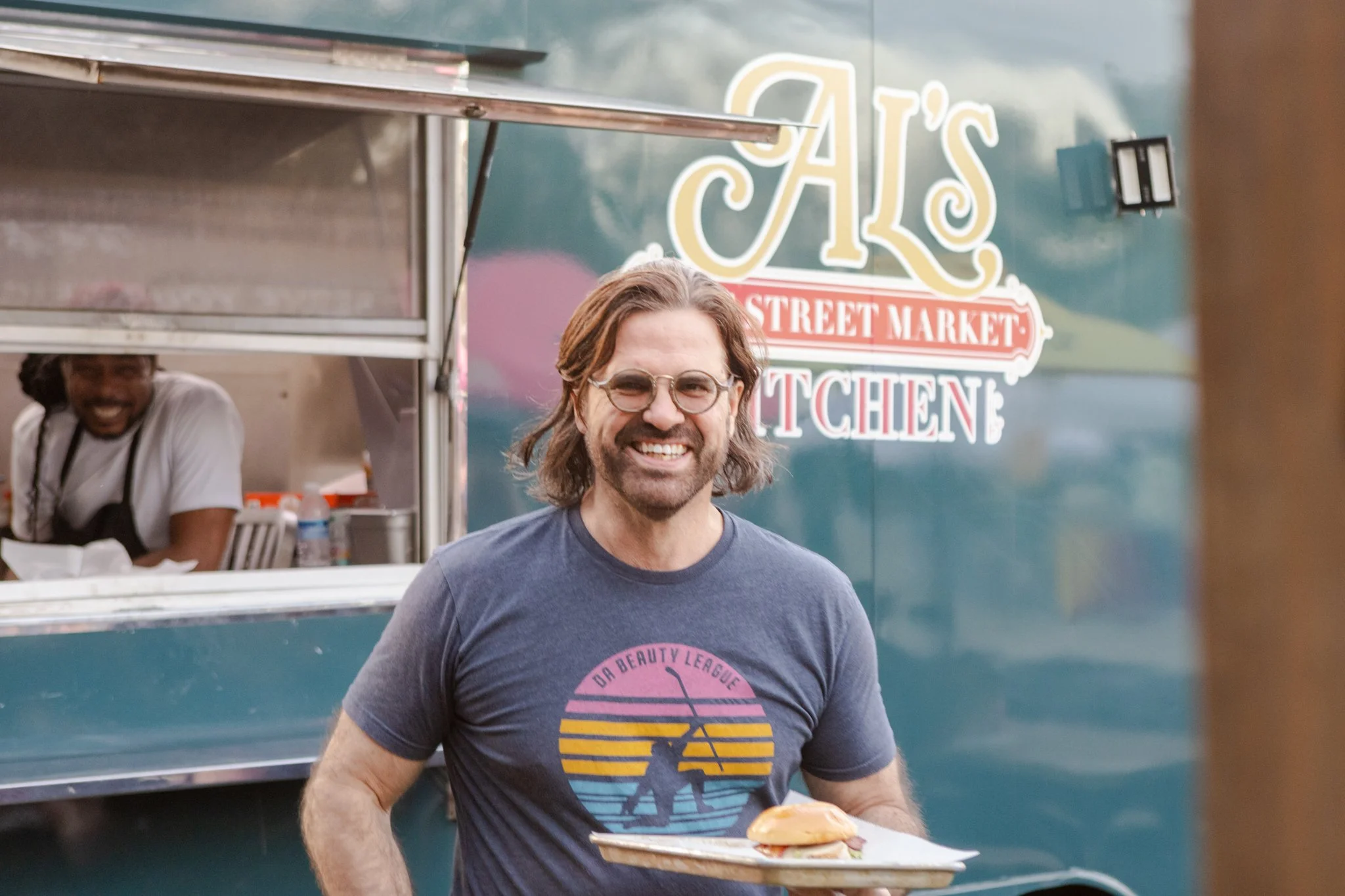 A man with glasses and long hair smiling while holding a plate with a sandwich in front of a food truck at a street market.