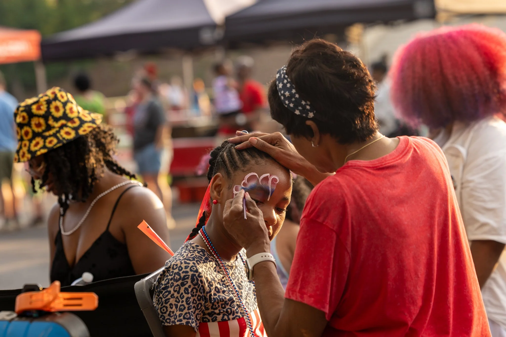 A woman is painting a child's face during an outdoor event, with other children and tents visible in the background.