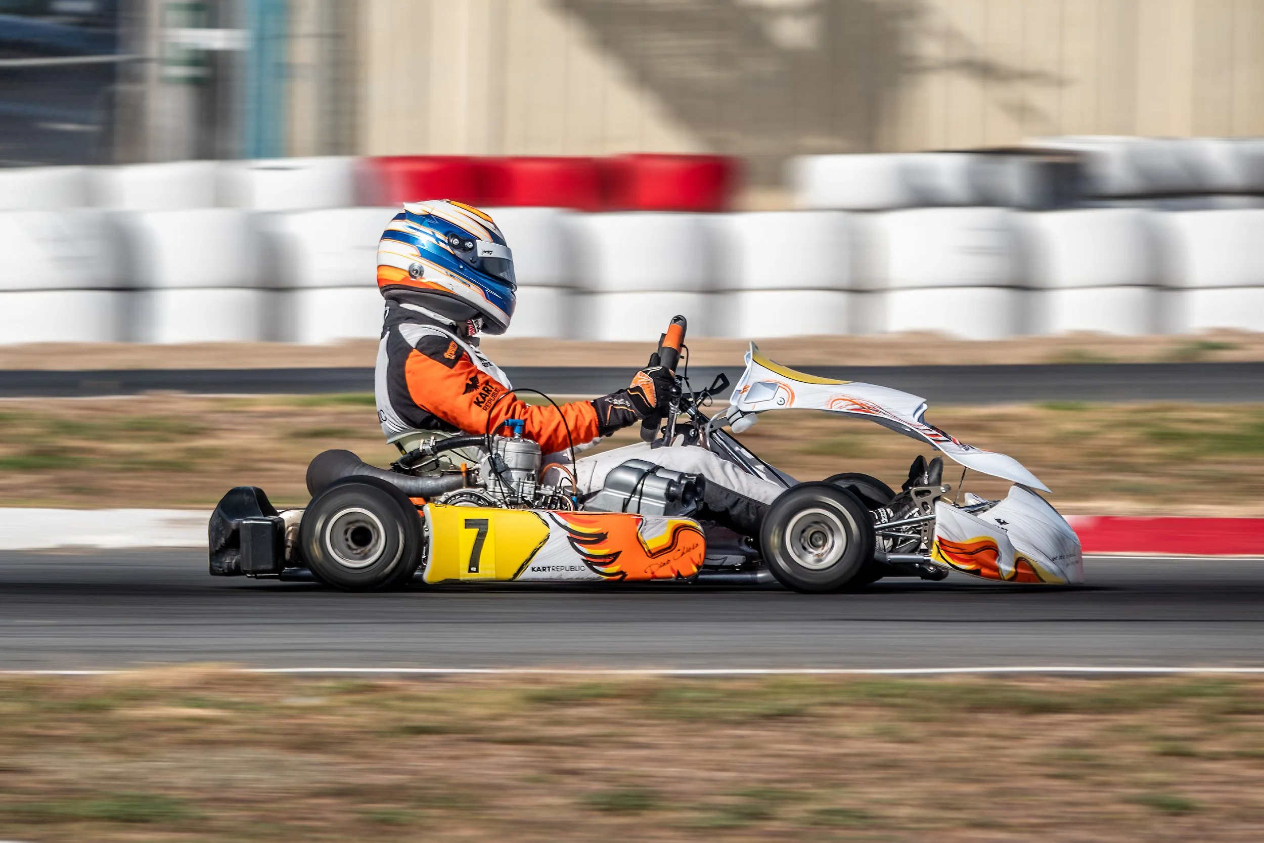 A go-kart with the number 7 being driven on a race track by a person in a racing suit and helmet, with a blurred background indicating high speed.