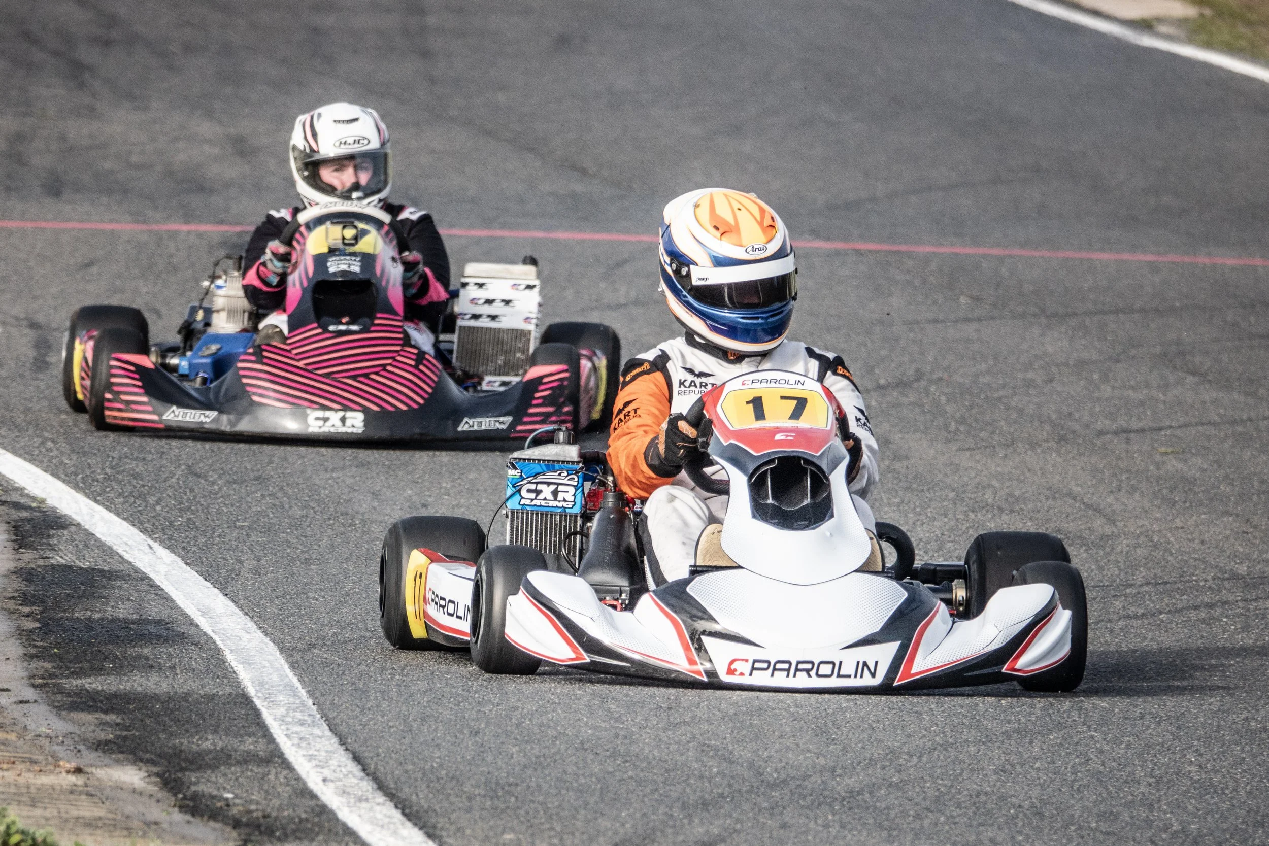 Two go-karts on a race track, driven by racers wearing helmets and racing suits.