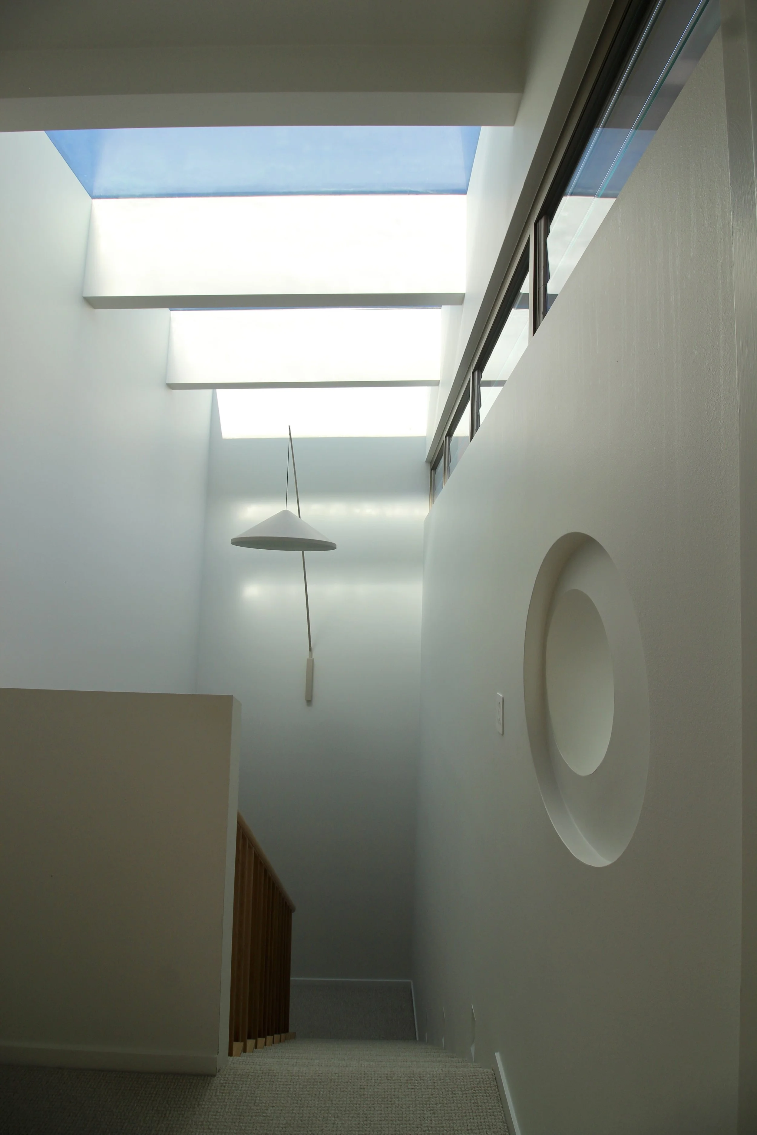 Interior view of a modern home with a high ceiling, large rectangular skylights, a hanging light fixture, a carpeted staircase, and a decorative wall feature with a circular indentation.
