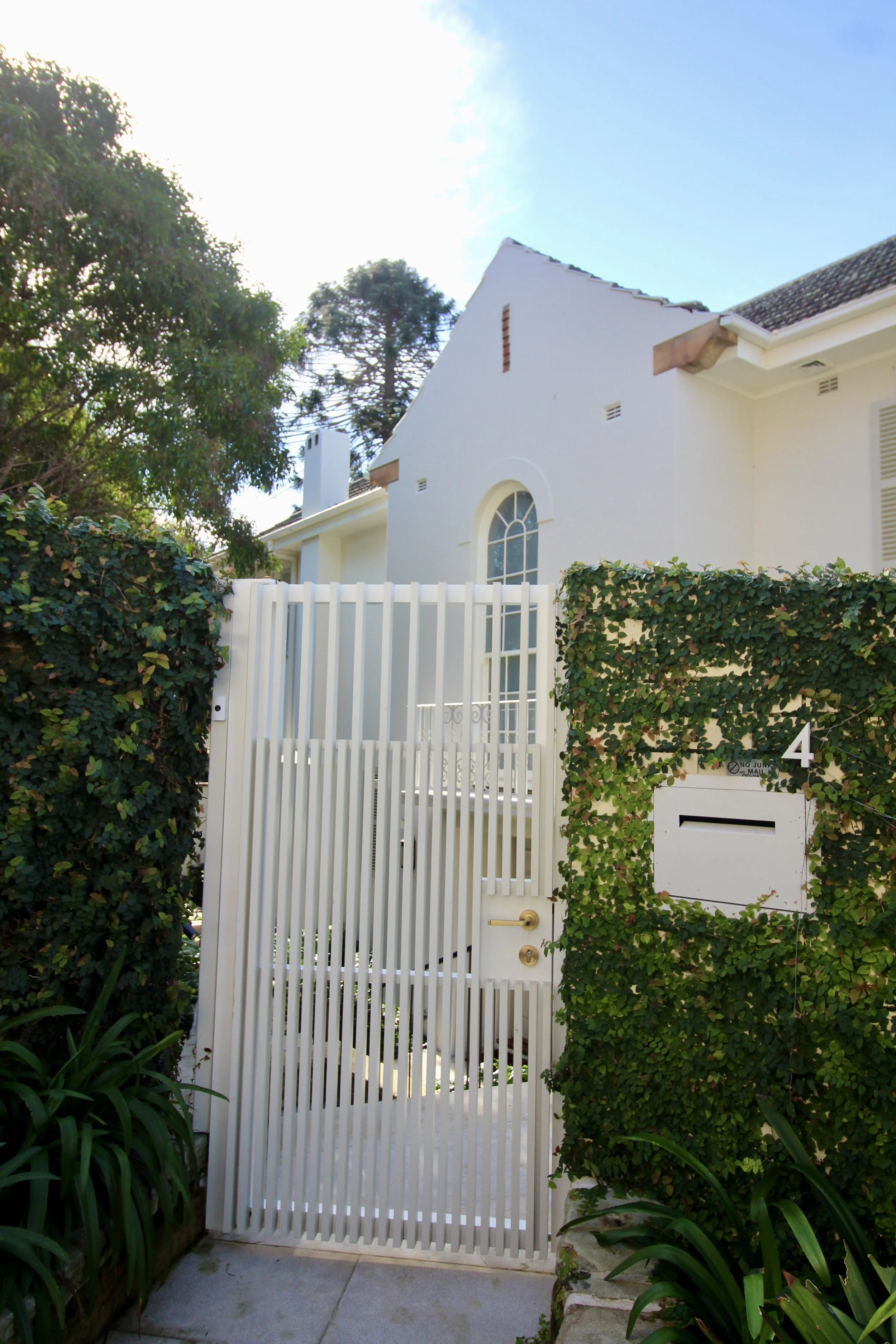 A white gate with vertical slats and a gold handle, surrounded by green ivy and plants, leading to a white house with a gabled roof, arched window, and brick accents, with trees and a blue sky in the background.