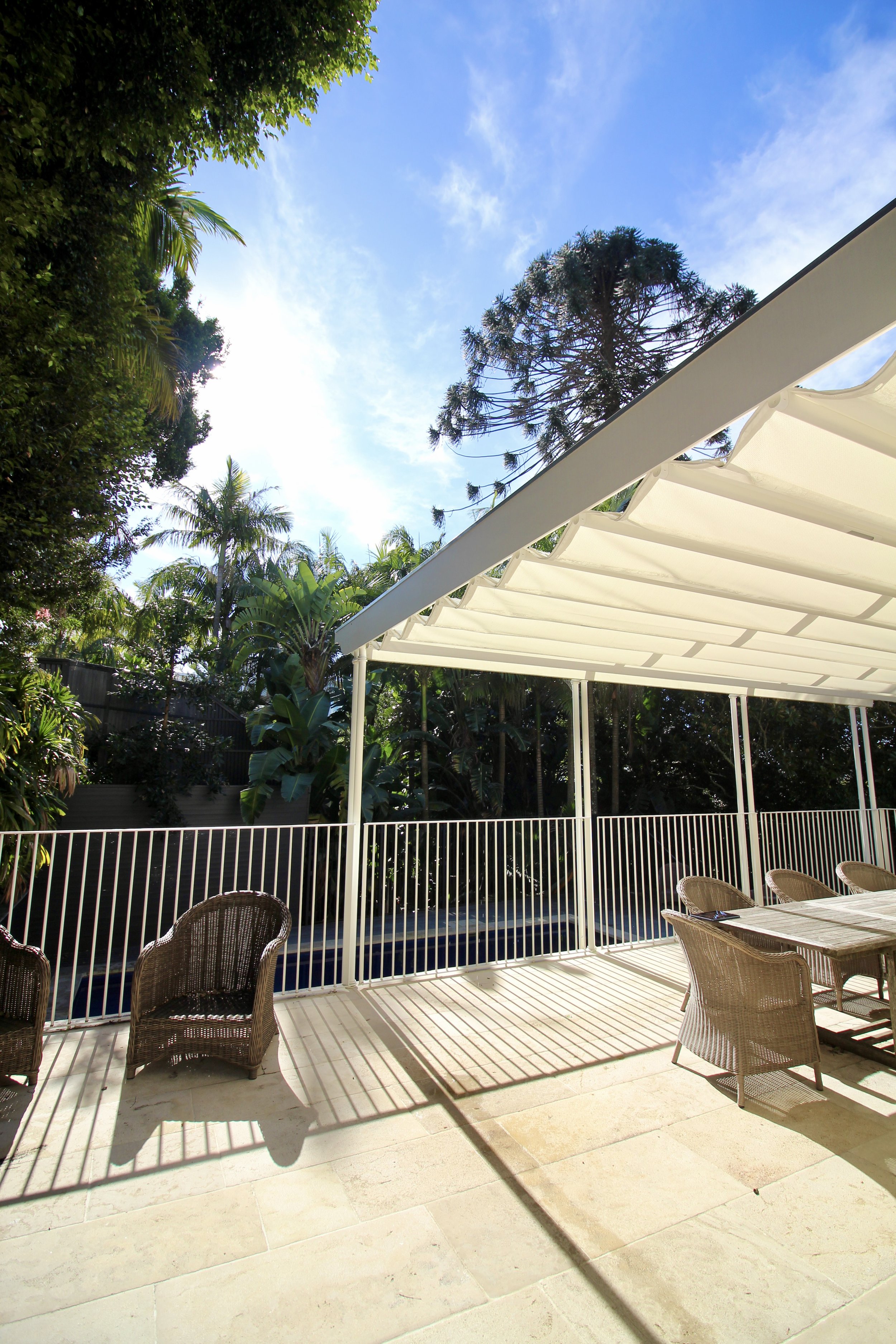 Outdoor patio area with wicker chairs, a dining table, and a white pergola, surrounded by lush green trees and plants under a clear blue sky.
