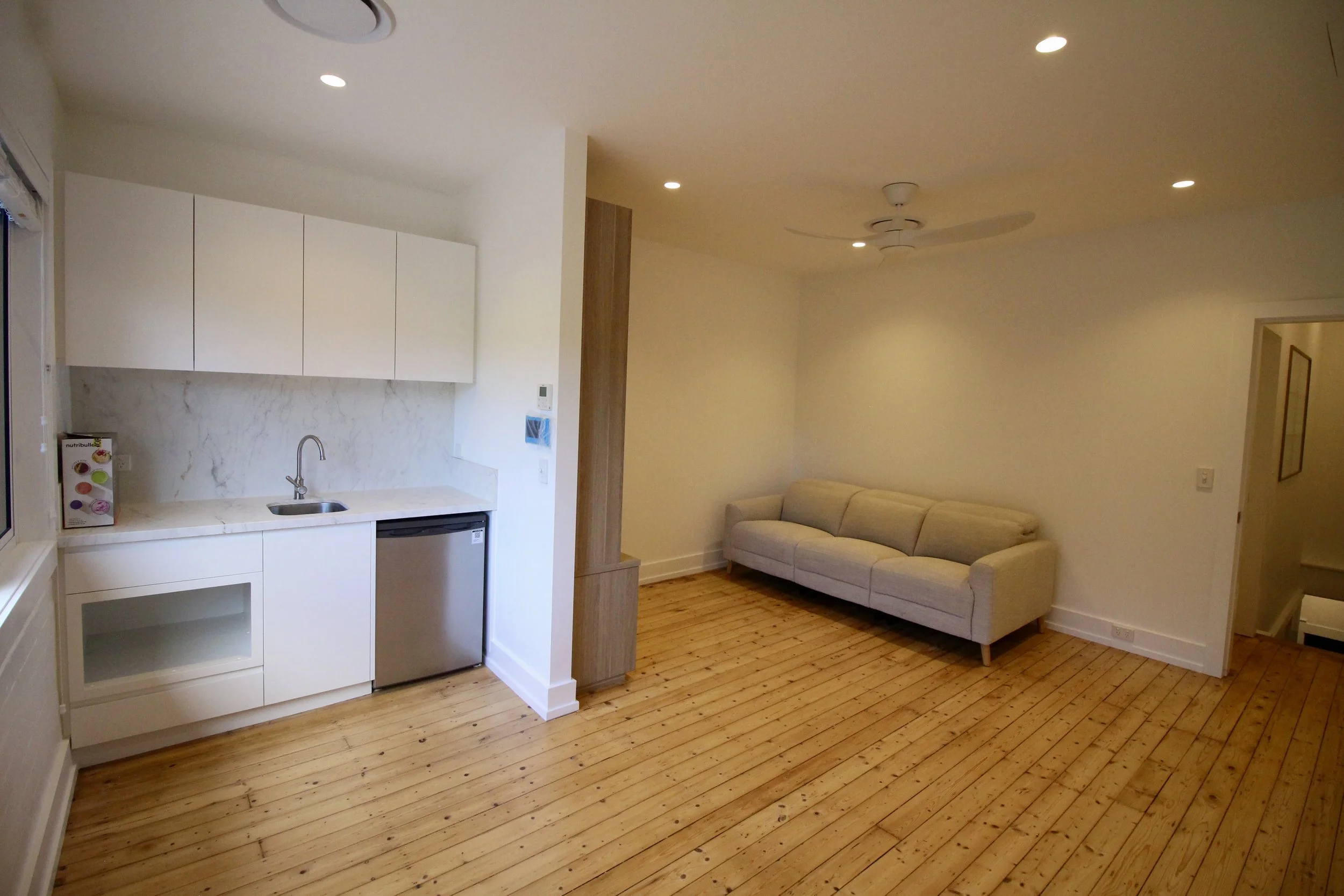 Interior of a modern apartment with a small kitchen area and a living room featuring a beige sofa, wooden floors, and recessed ceiling lights.