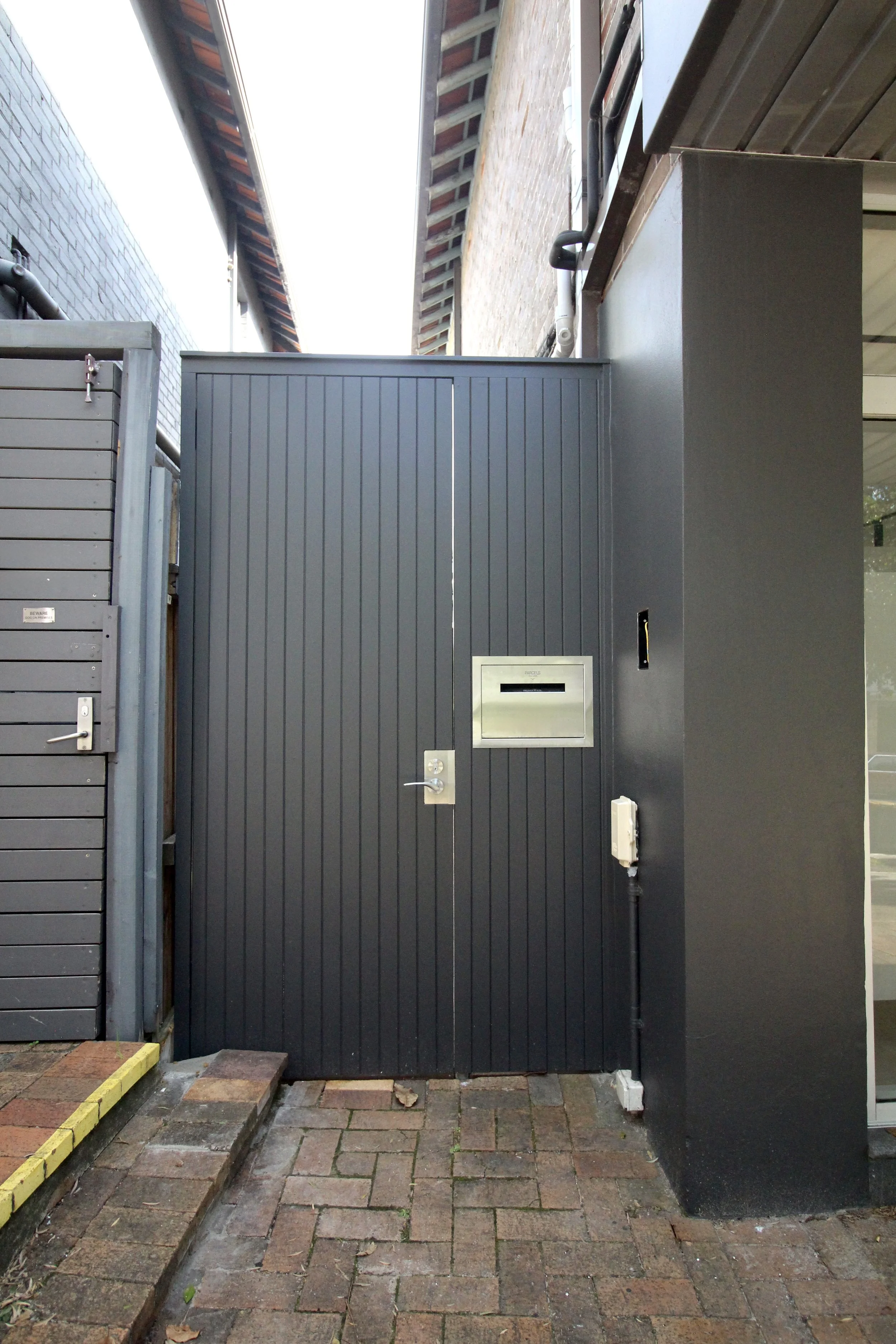 A black metal garage door with a mailbox and an intercom system mounted on it, in an alleyway with brick paving and a yellow border on the left side.