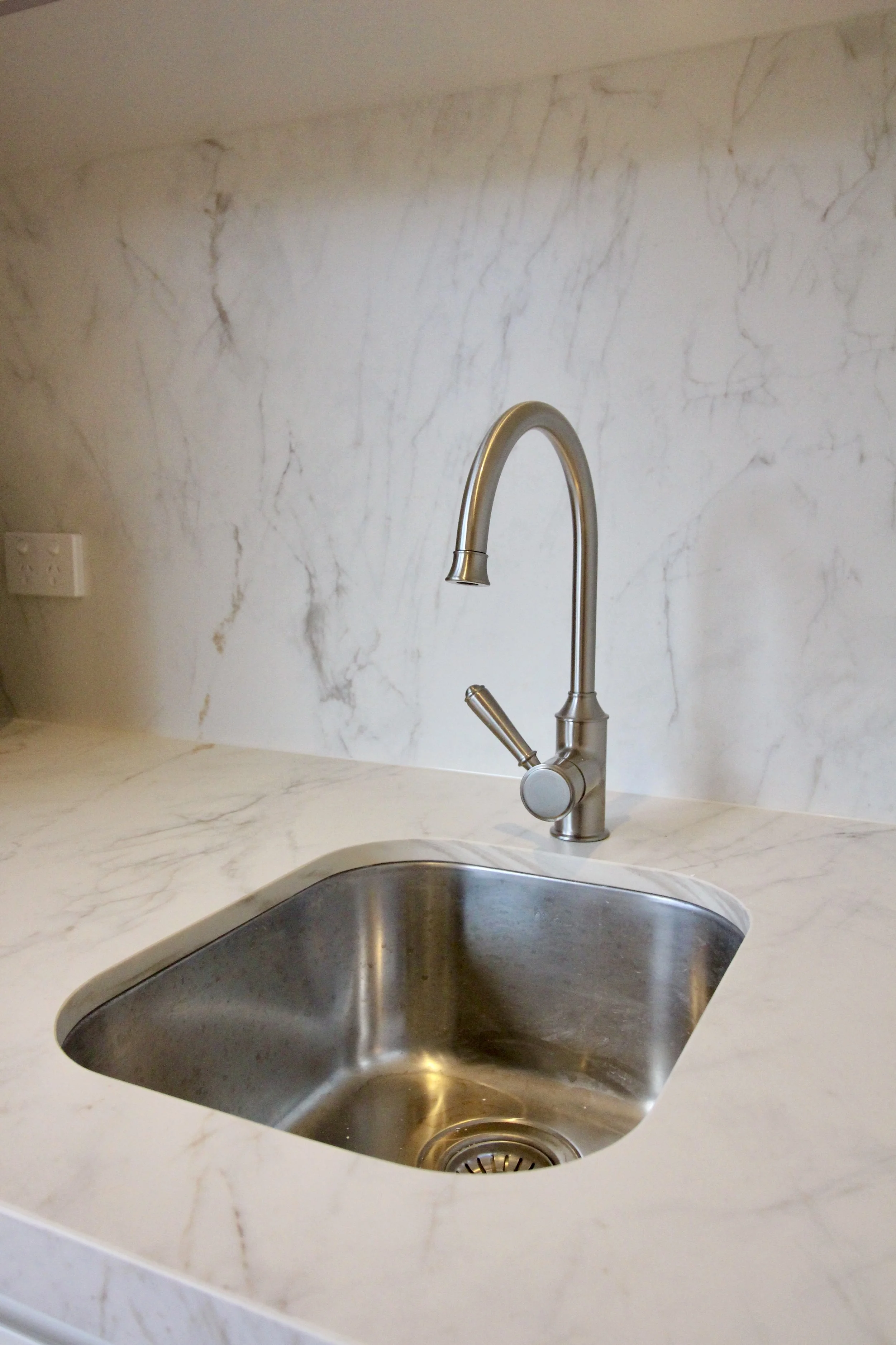 Kitchen sink with stainless steel basin and a modern curved faucet installed in a white marble countertop with gray veining and a matching marble backsplash. An electrical outlet is visible on the wall behind the sink.