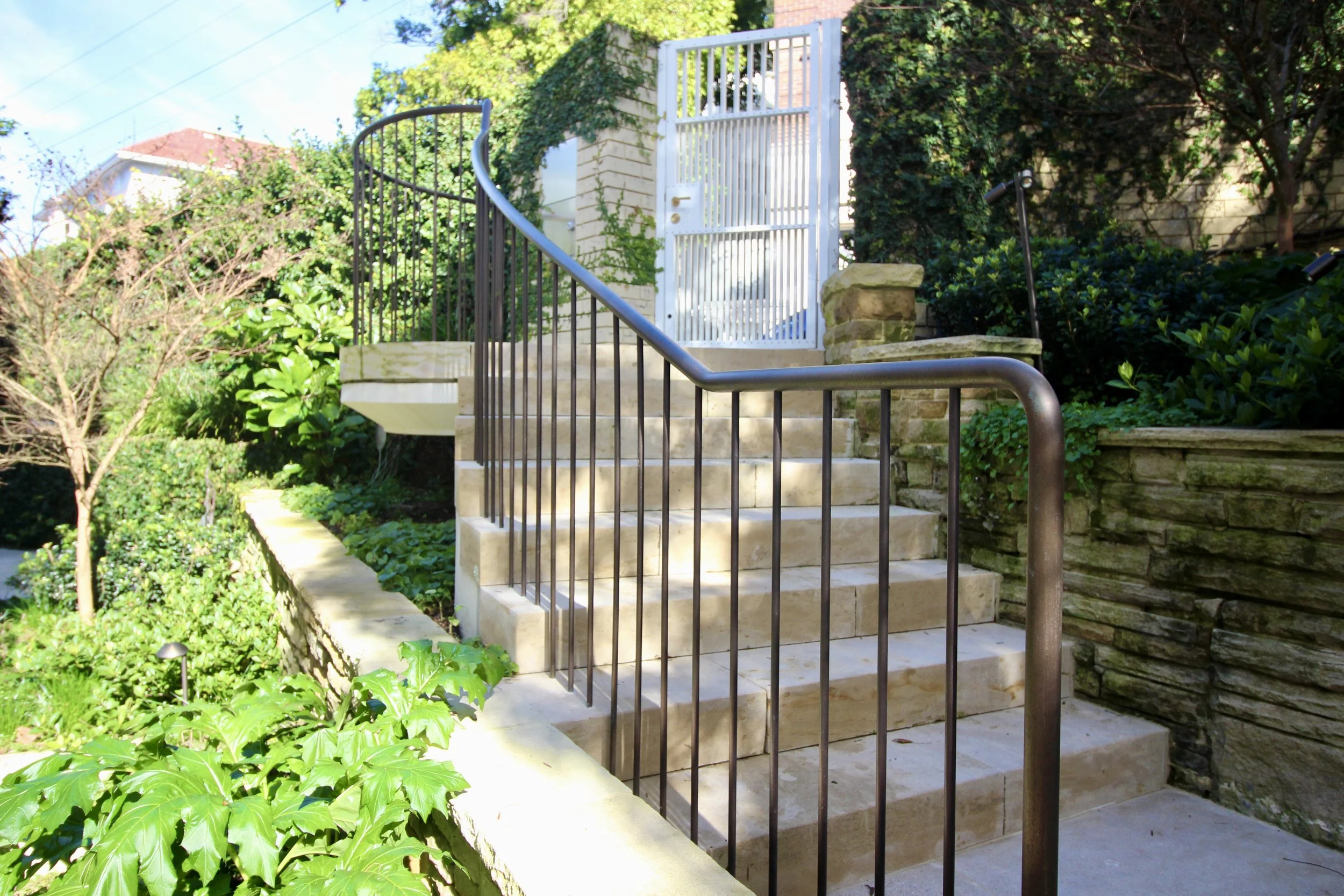 Exterior staircase with beige stone steps and black metal railing, leading to a white metal gate surrounded by lush green plants and bushes, with a brick wall in the background.