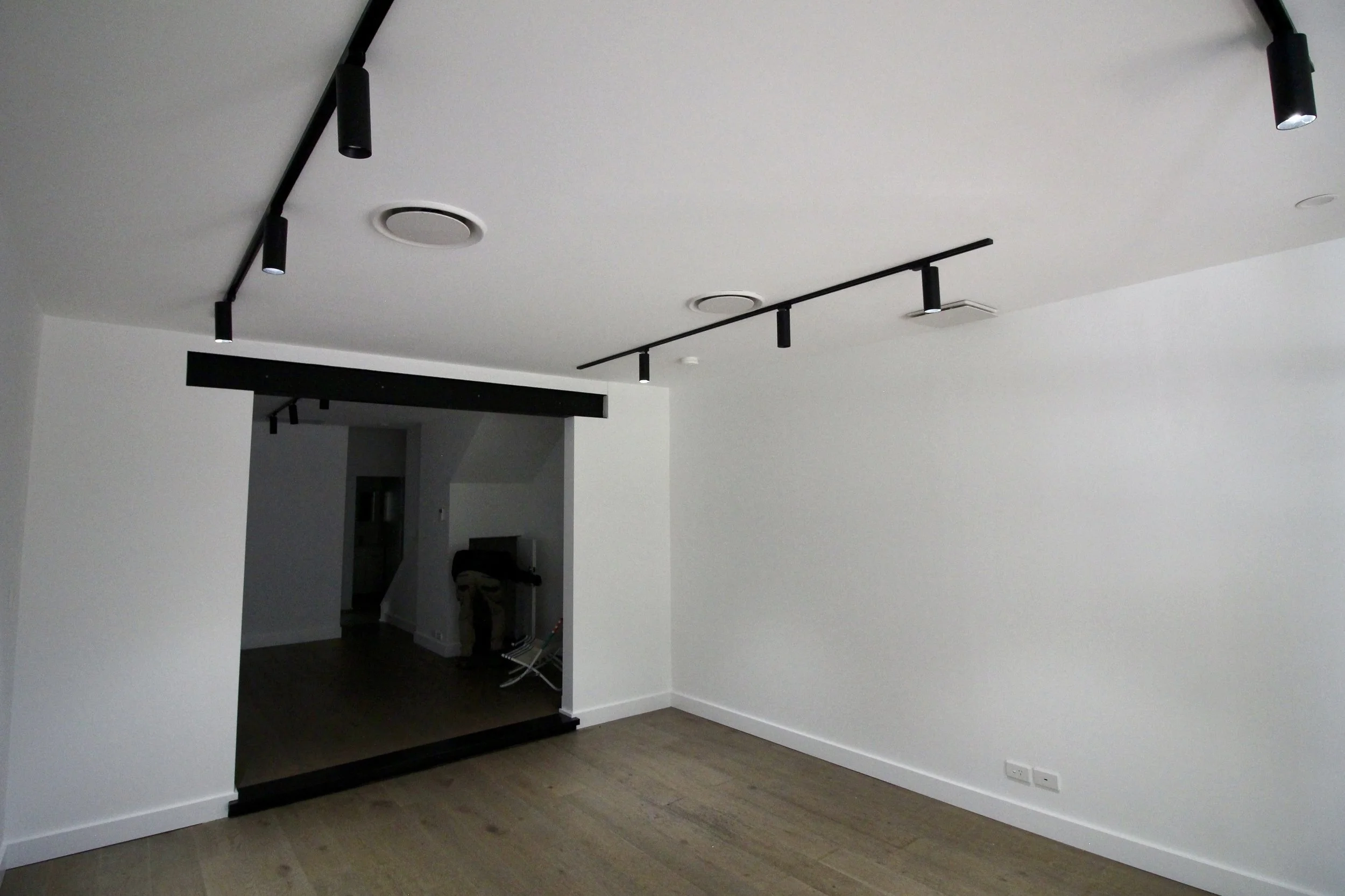 Interior of a room with white walls and ceiling, featuring black track lighting, a black ceiling-mounted structure, and a light-colored wooden floor.