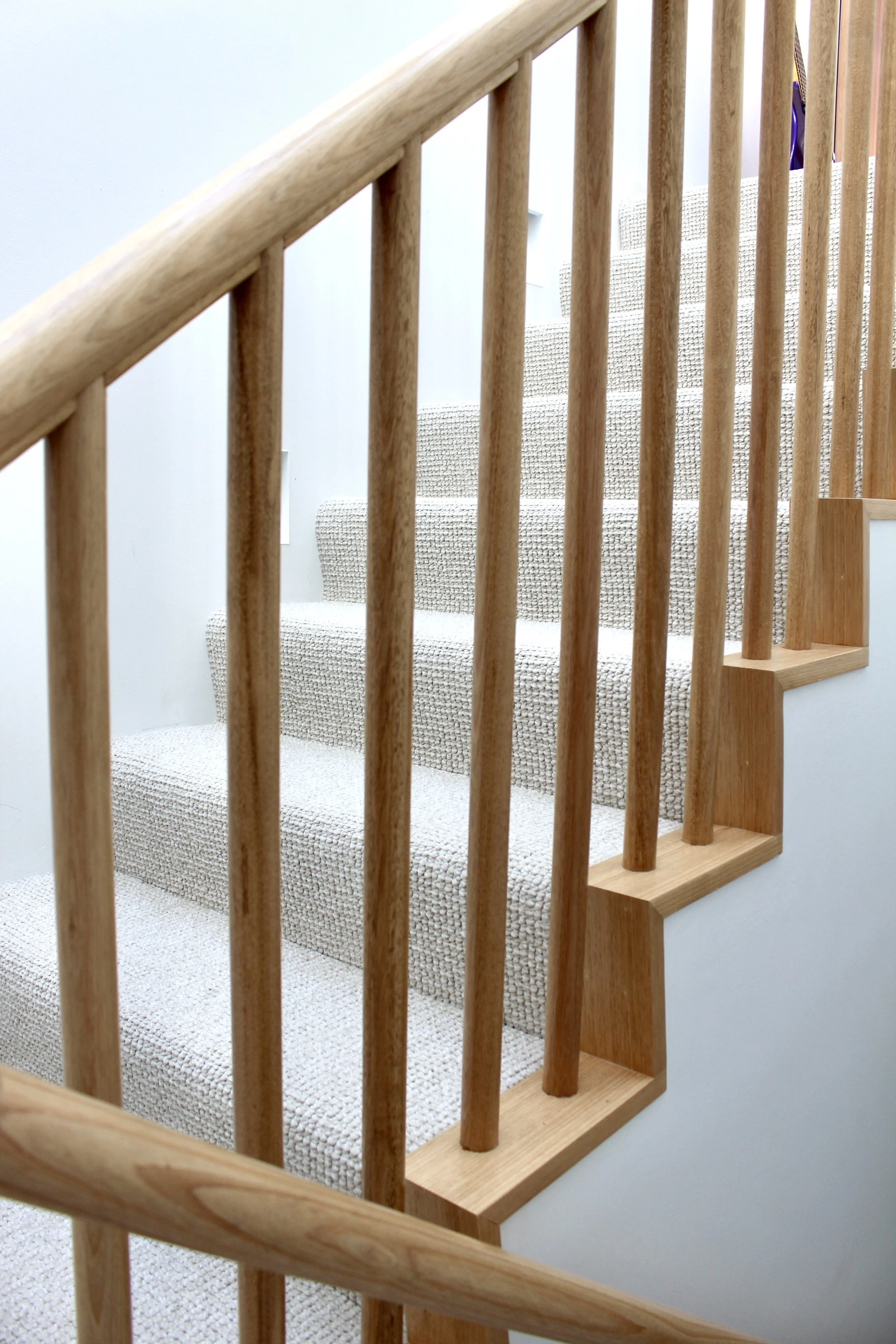 Wooden staircase with banister and spindle railing, beige carpeted steps against a white wall.