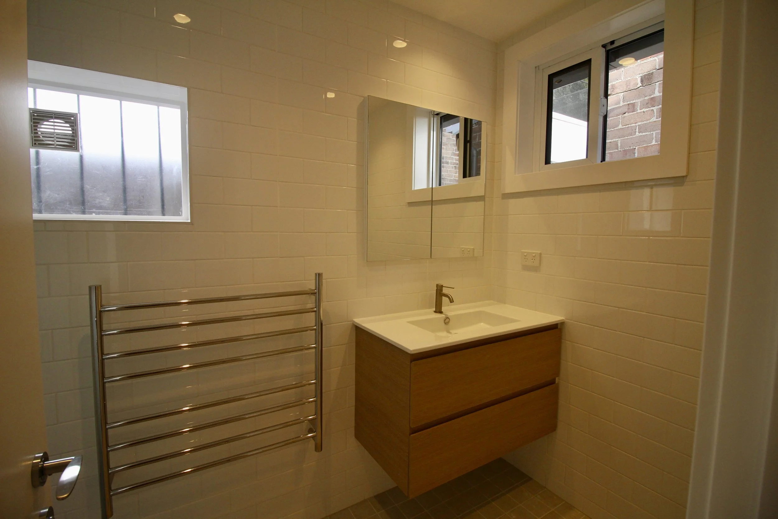 A modern bathroom with beige tiled walls, a small white sink with vanity, a mirror, a heated towel rail, and two windows showing brick exterior walls outside.