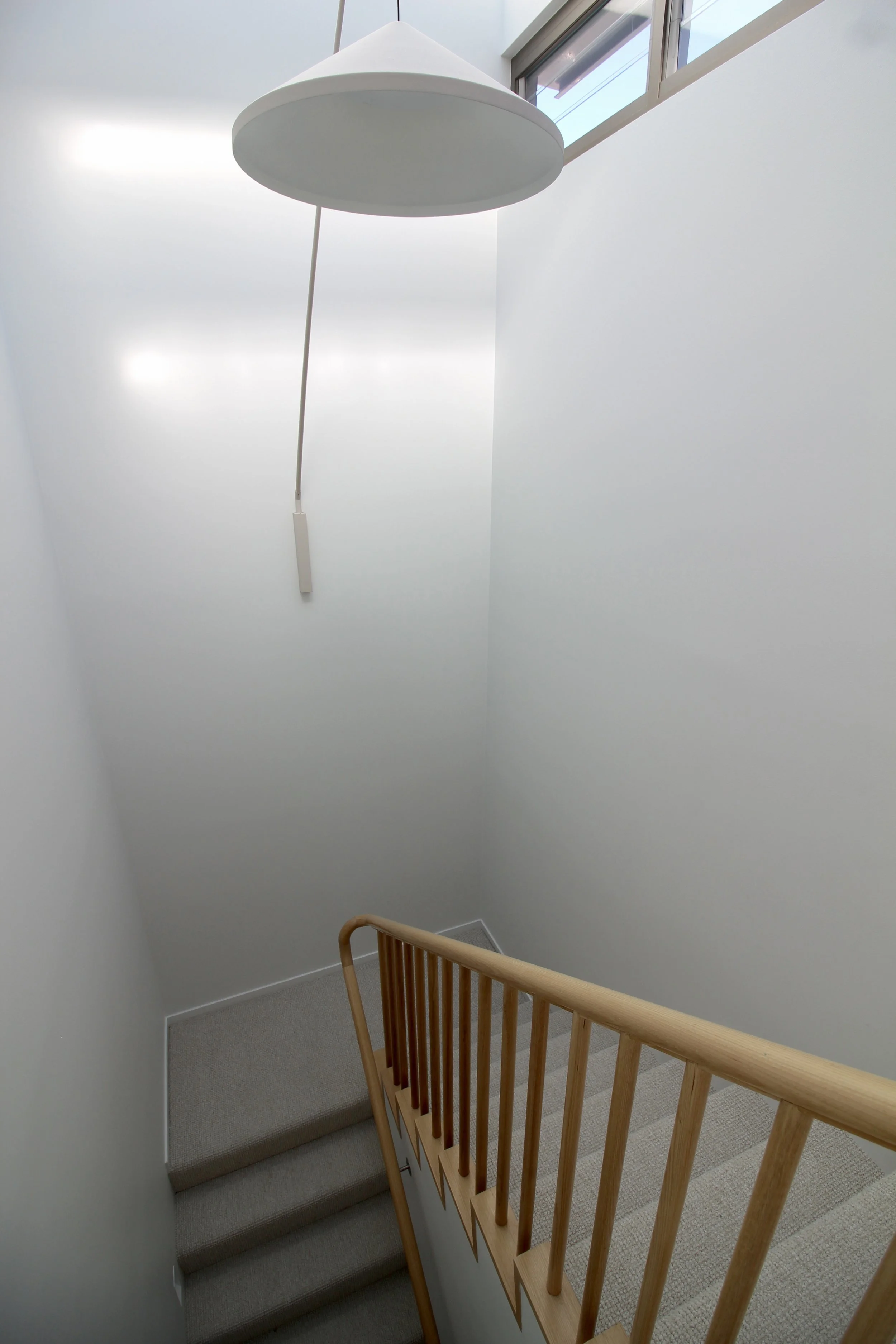 A staircase with beige carpeting, light-colored wooden handrails, and white walls. A window near the ceiling lets natural light into the space.