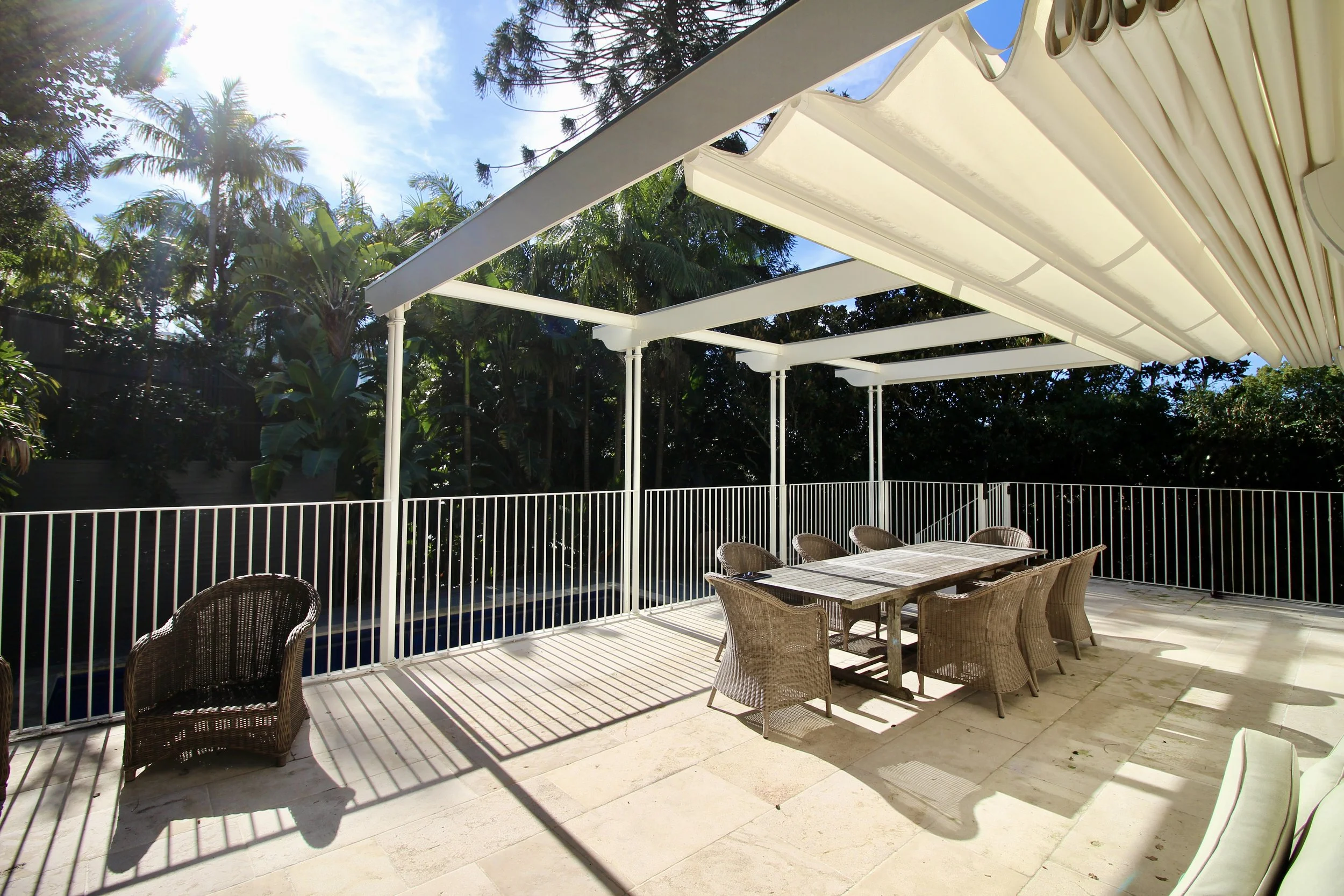 A patio with a large dining table and wicker chairs under a white retractable awning, surrounded by lush greenery and trees, with sunlight shining through.