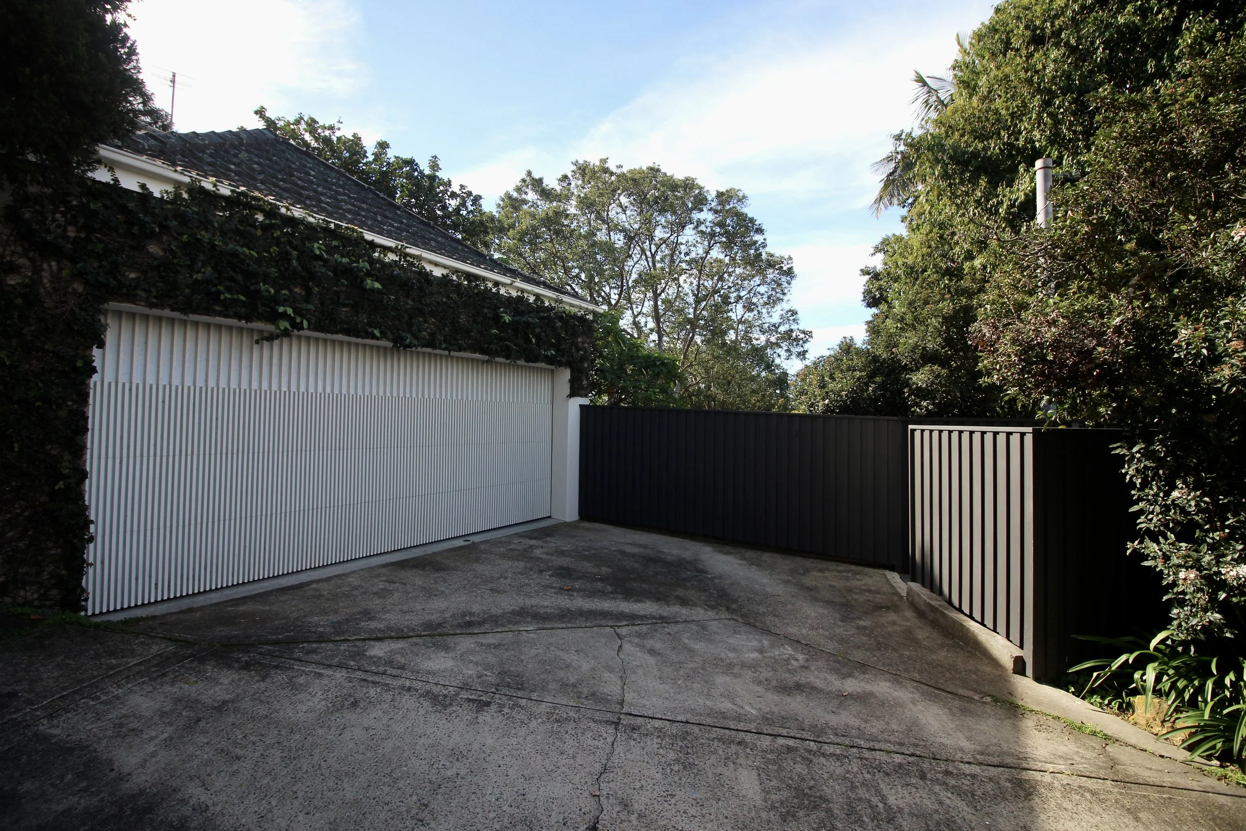 A residential driveway with a white garage door and a black side gate, surrounded by green trees and bushes under a blue sky.