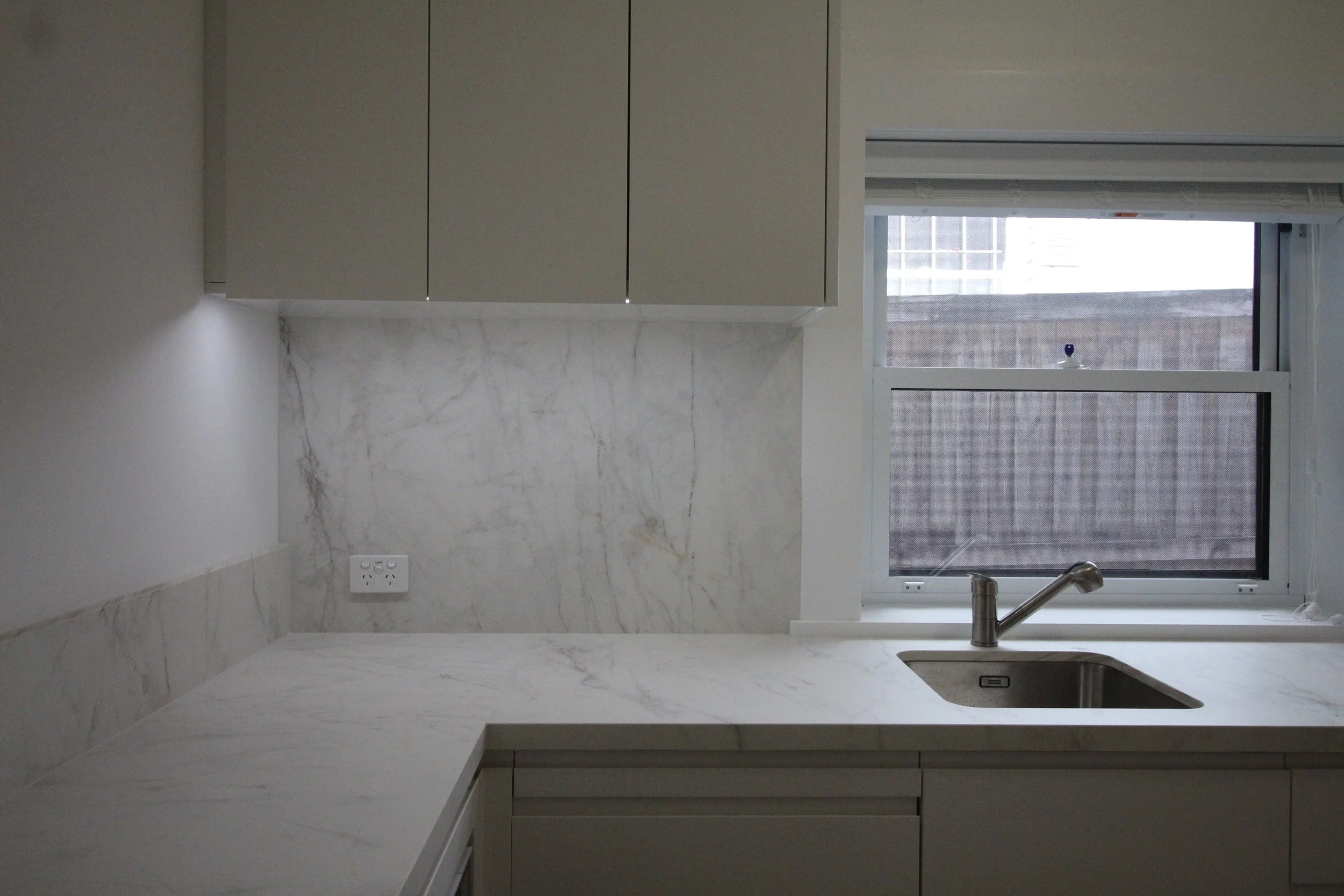 A modern kitchen with white marble countertops, a stainless steel faucet, and a window with a view of an outdoor fence.