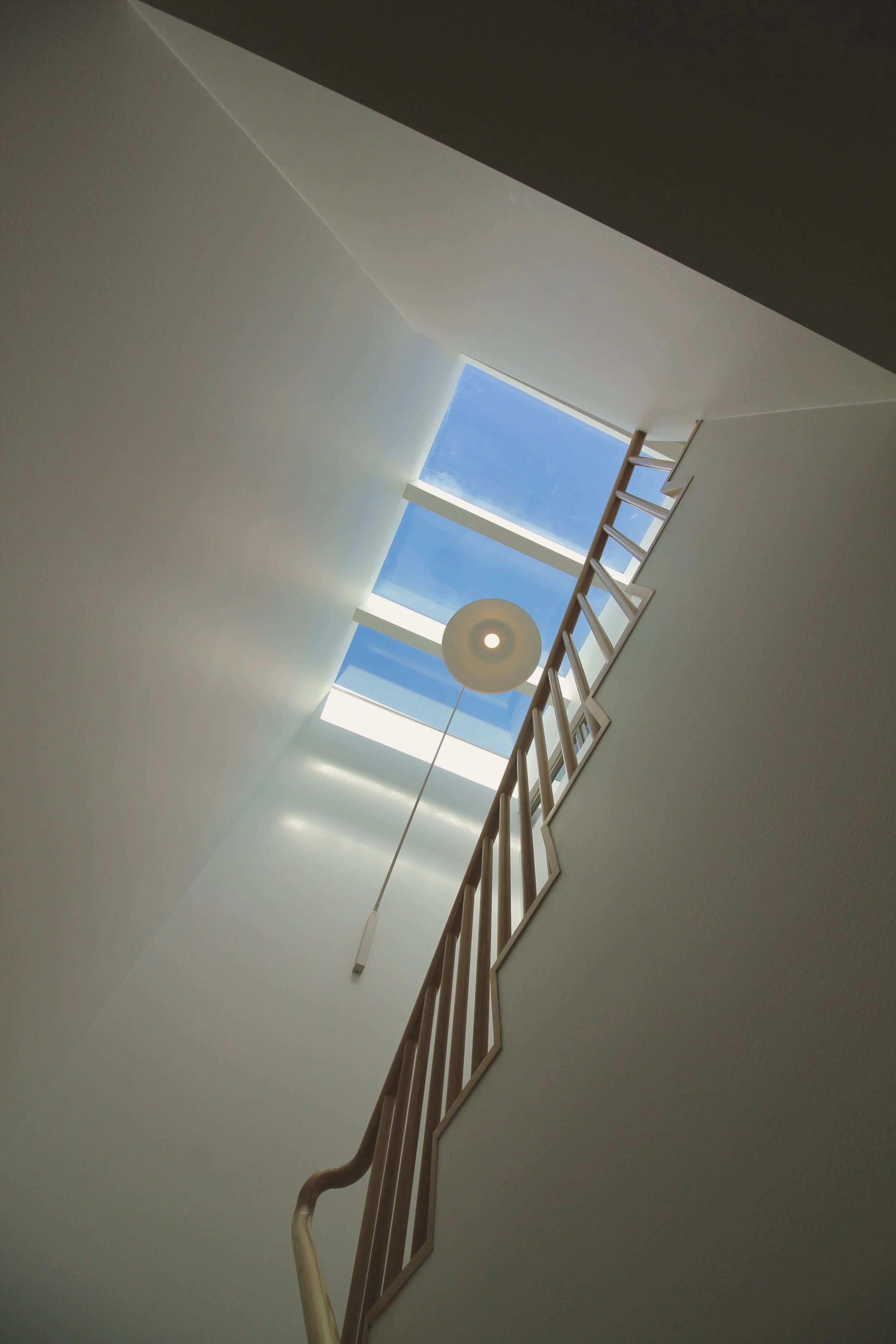 Interior shot of a staircase viewed from below, with a skylight at the top showing blue sky and white clouds, a wooden handrail, and an overhead ceiling light.