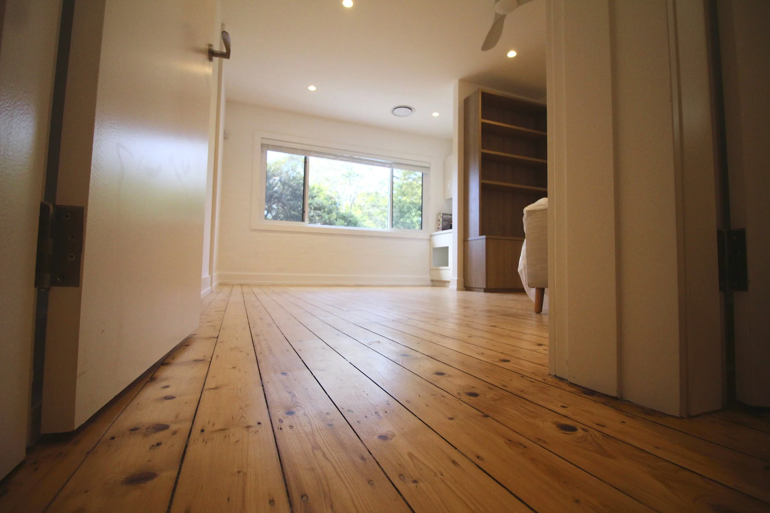 A room with wooden flooring, white walls, a window showing green trees outside, a bookshelf, and part of a chair.