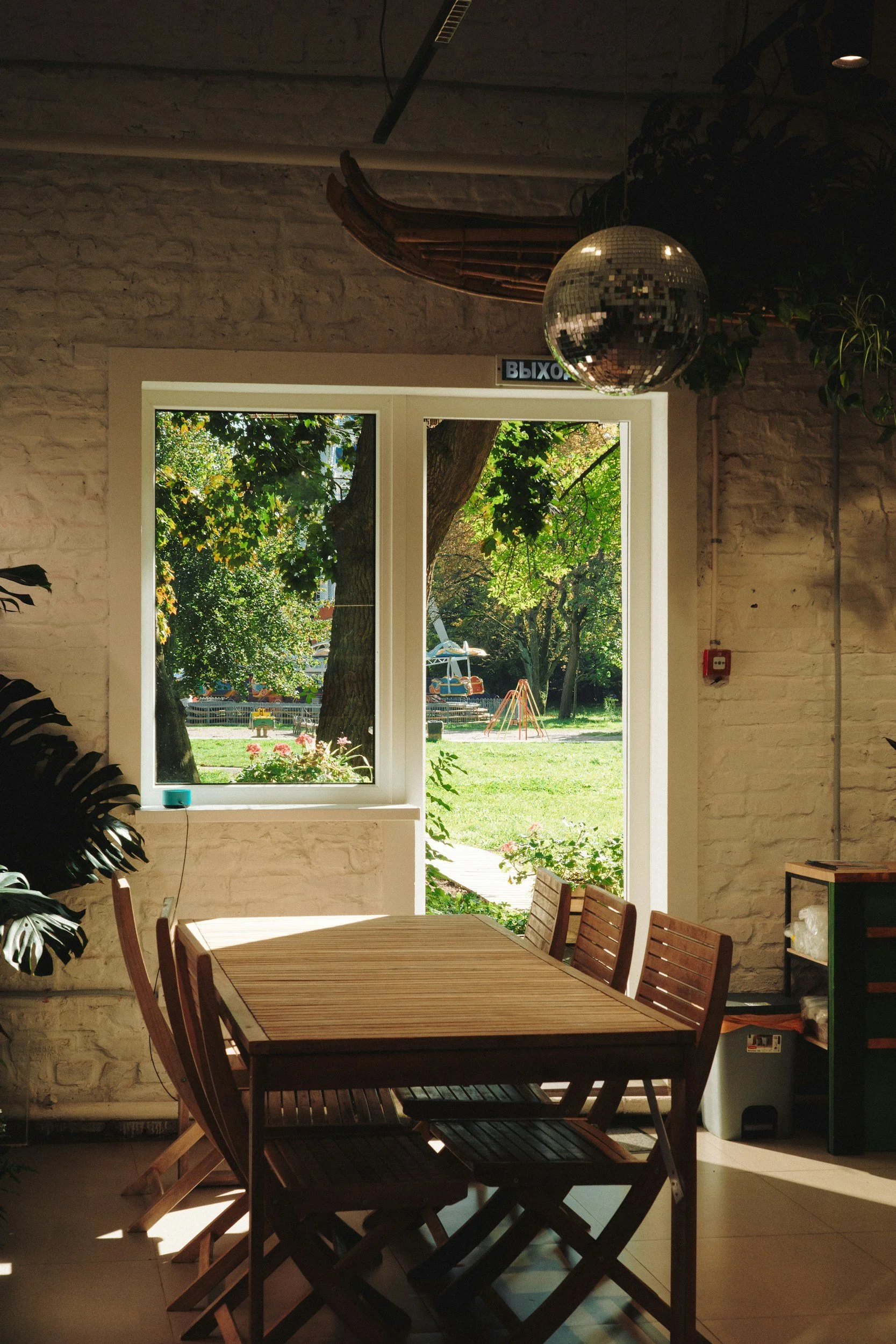 Interior of a room with a wooden dining table and chairs, a large window showing greenery and a playground outside, a disco ball hanging from the ceiling, and a white brick wall.
