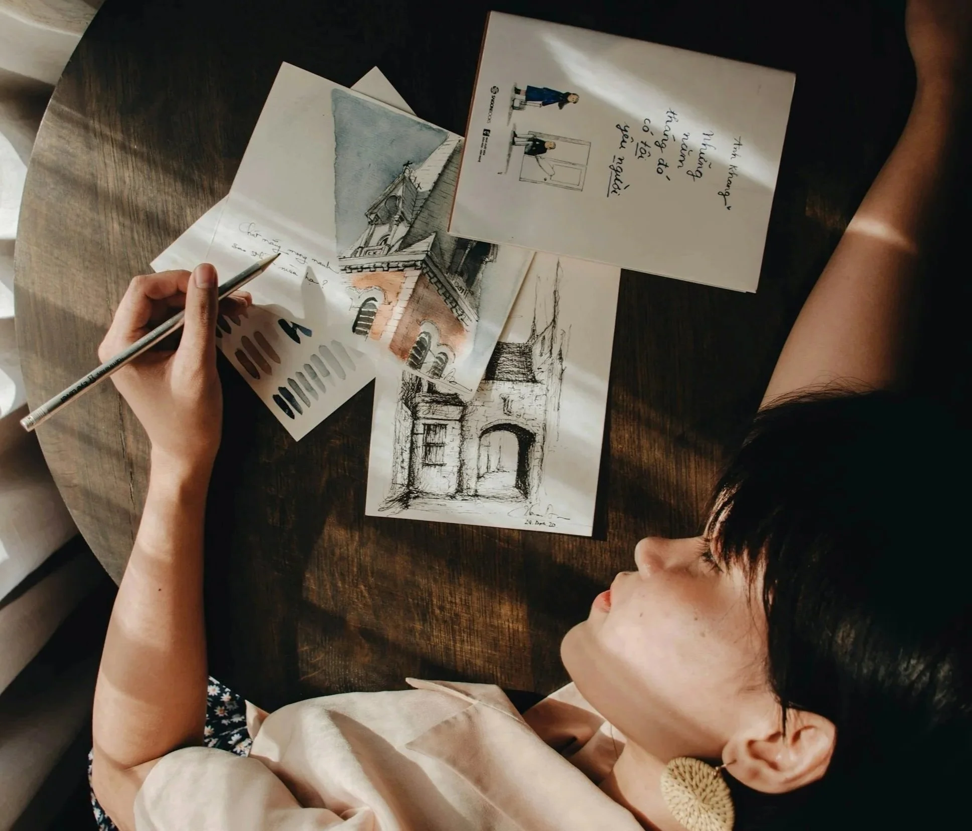 A person is sitting at a wooden table, drawing and painting architectural buildings on paper with colored pencils. There are multiple sketch drawings and watercolor paintings of buildings, including a church and other structures, spread out on the table. The scene is well-lit.