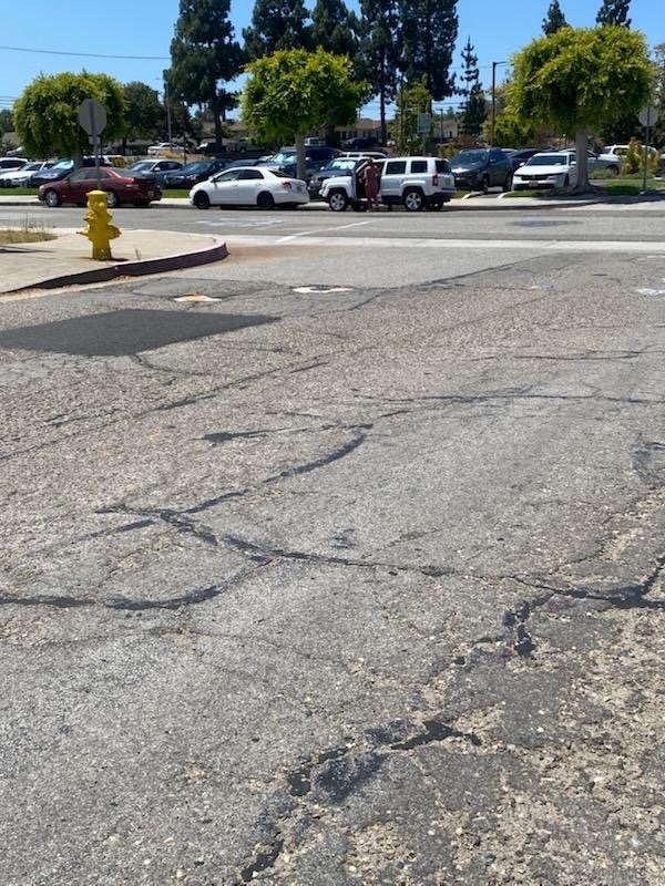 A street with cracked asphalt in the foreground, a yellow fire hydrant on the sidewalk, and a parking lot with several parked cars and trees in the background.