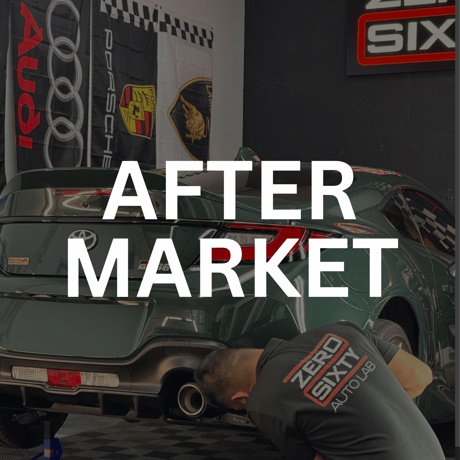 A green Toyota Supra in an auto workshop, with a mechanic working underneath it. The workshop has banners of Porsche, Audi, and Lamborghini, and a sign that reads 'SIX' in the background.