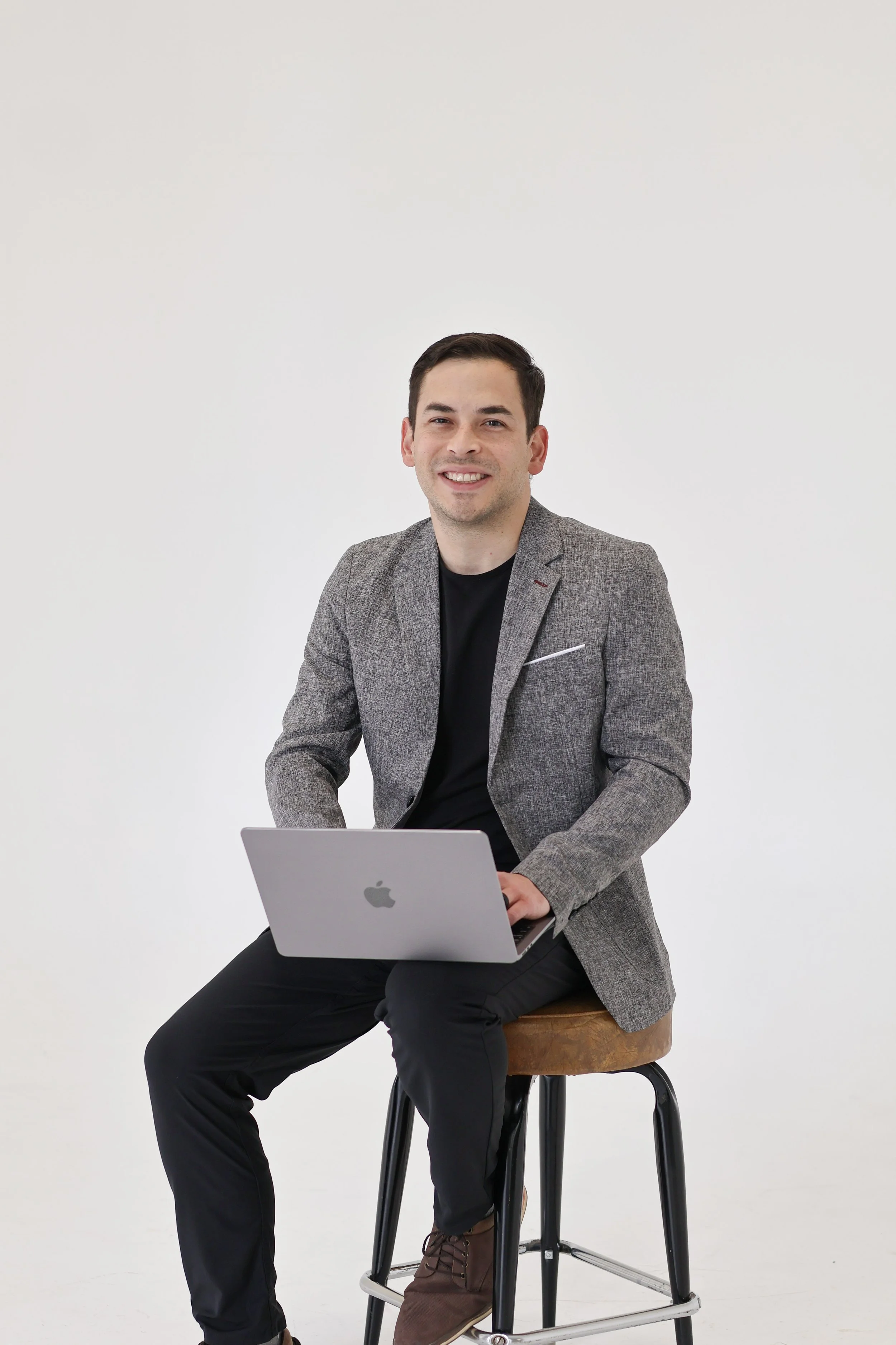 A man with short dark hair sitting on a high stool, smiling, using a silver Apple MacBook laptop on his lap, dressed in a gray blazer, black shirt, and black pants, against a plain white background.