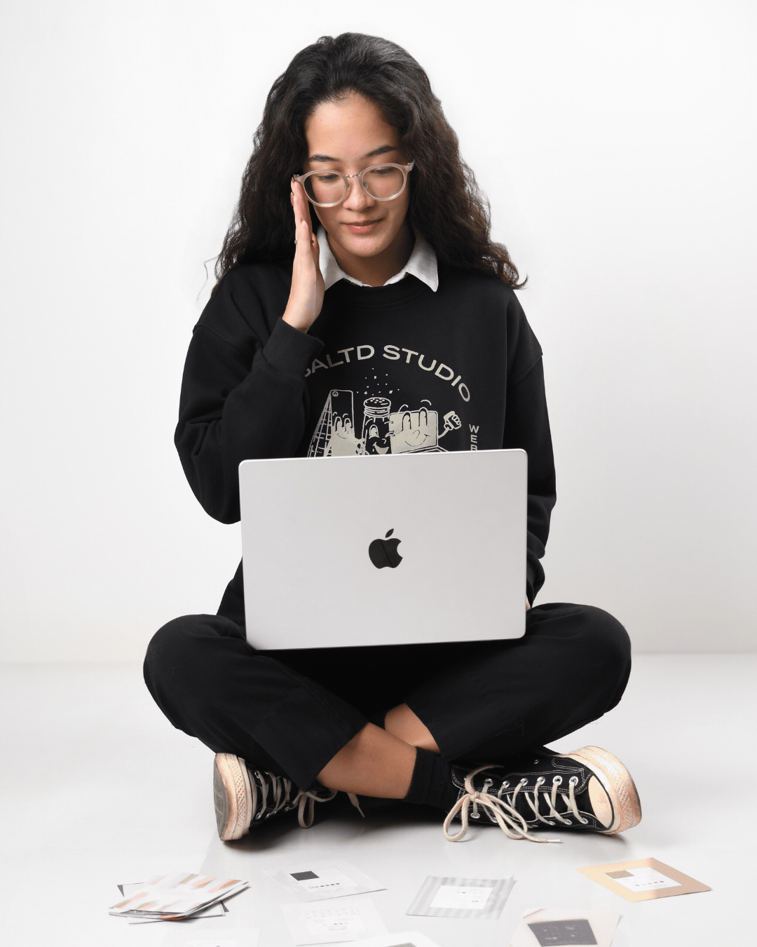 A young woman sitting cross-legged on the floor, using a silver MacBook, with her right hand touching her face. She is wearing glasses, a black sweatshirt with graphics, black pants, and black sneakers with white laces and soles. There are printed photo cards spread on the floor in front of her, and she appears to be working or reviewing something on her laptop.