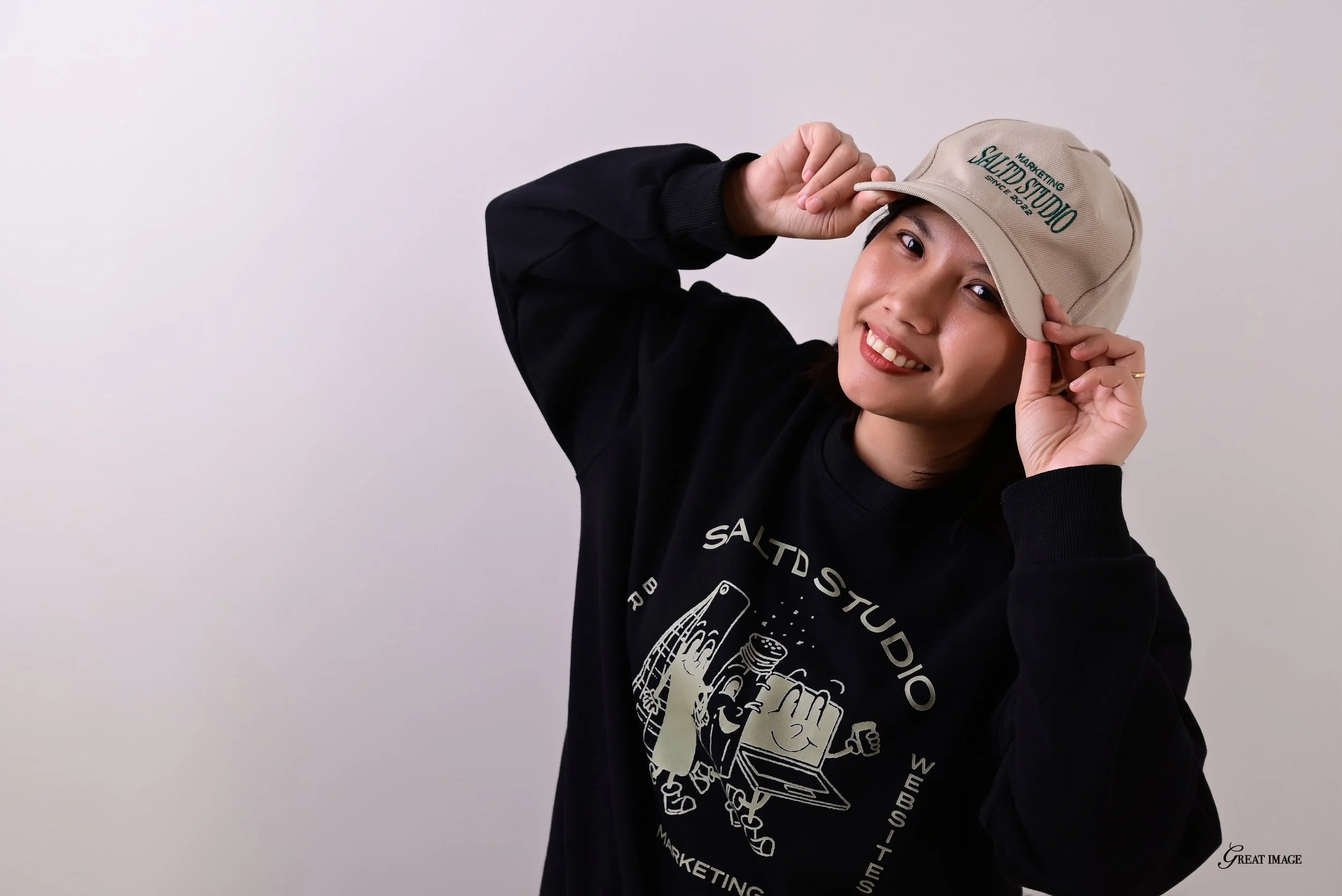 Young woman smiling, wearing a black sweatshirt and a beige cap with green text, against a plain light background.