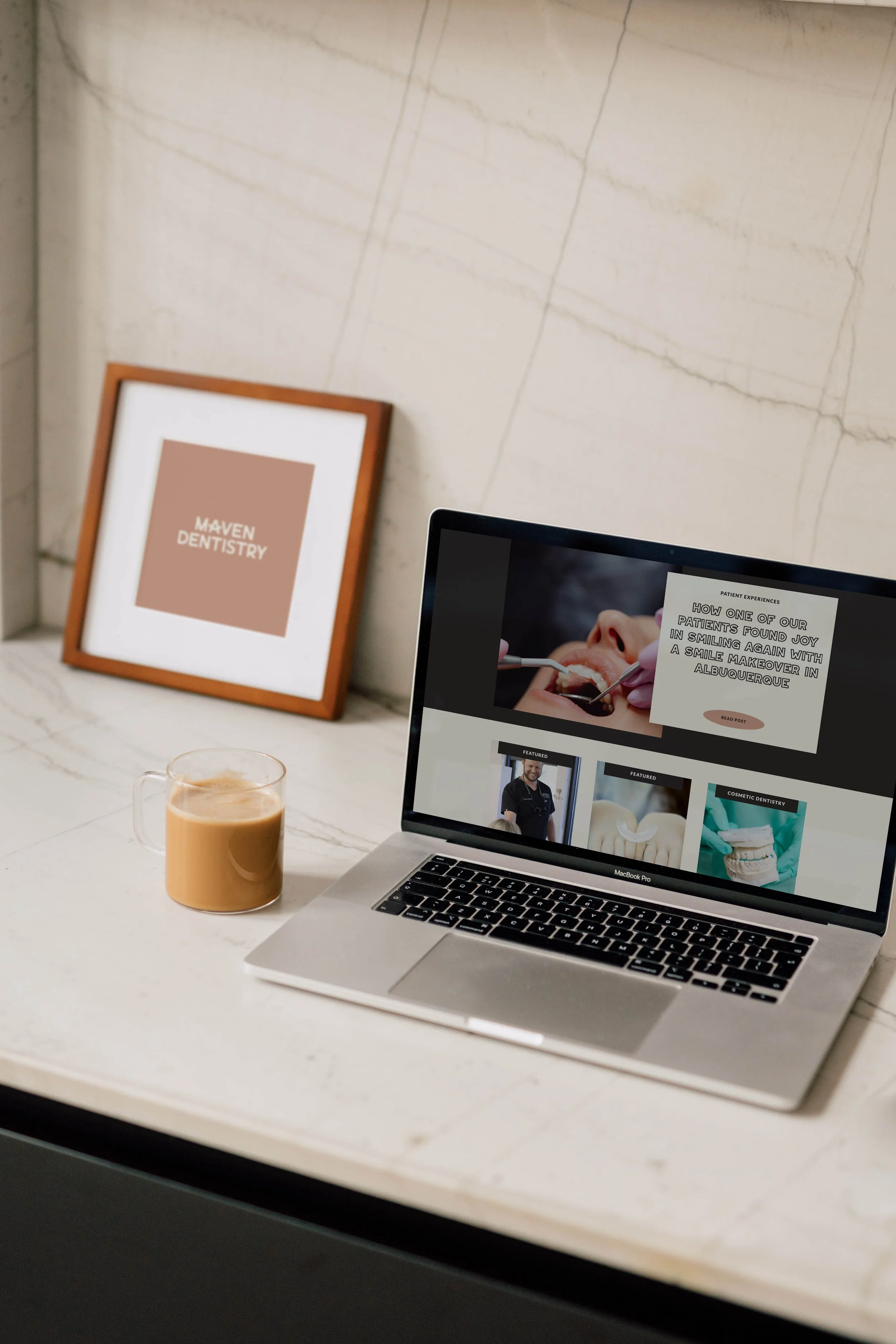 A laptop on a white marble desk displaying a website about dentistry, with a coffee mug beside it, and framed artwork on the wall that says "Maven Dentistry."