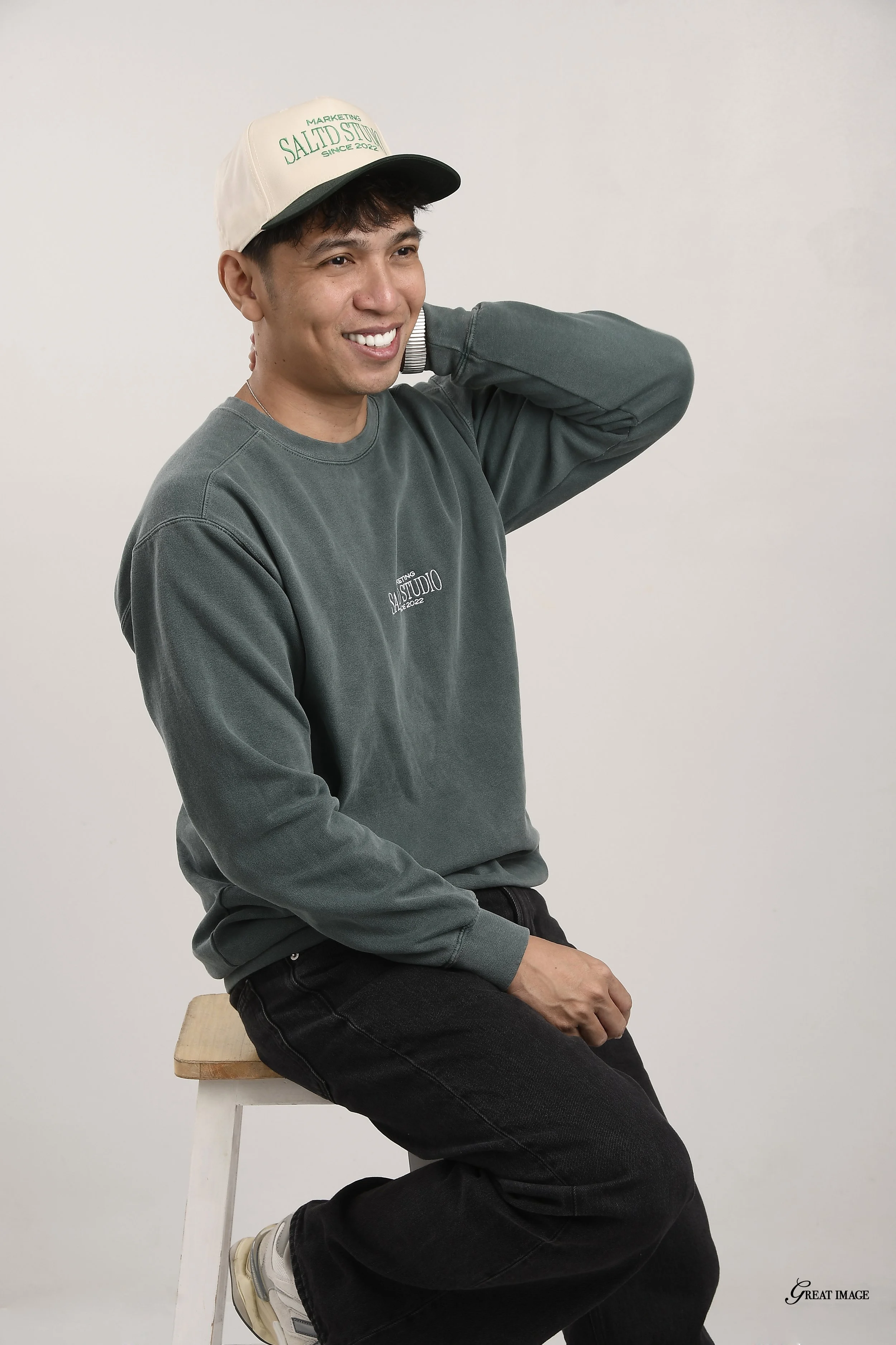 A smiling young man with short dark hair, wearing a beige and black baseball cap, gray sweatshirt with some writing, and black jeans, sitting on a wooden stool against a plain white background, with his right hand resting behind his head.