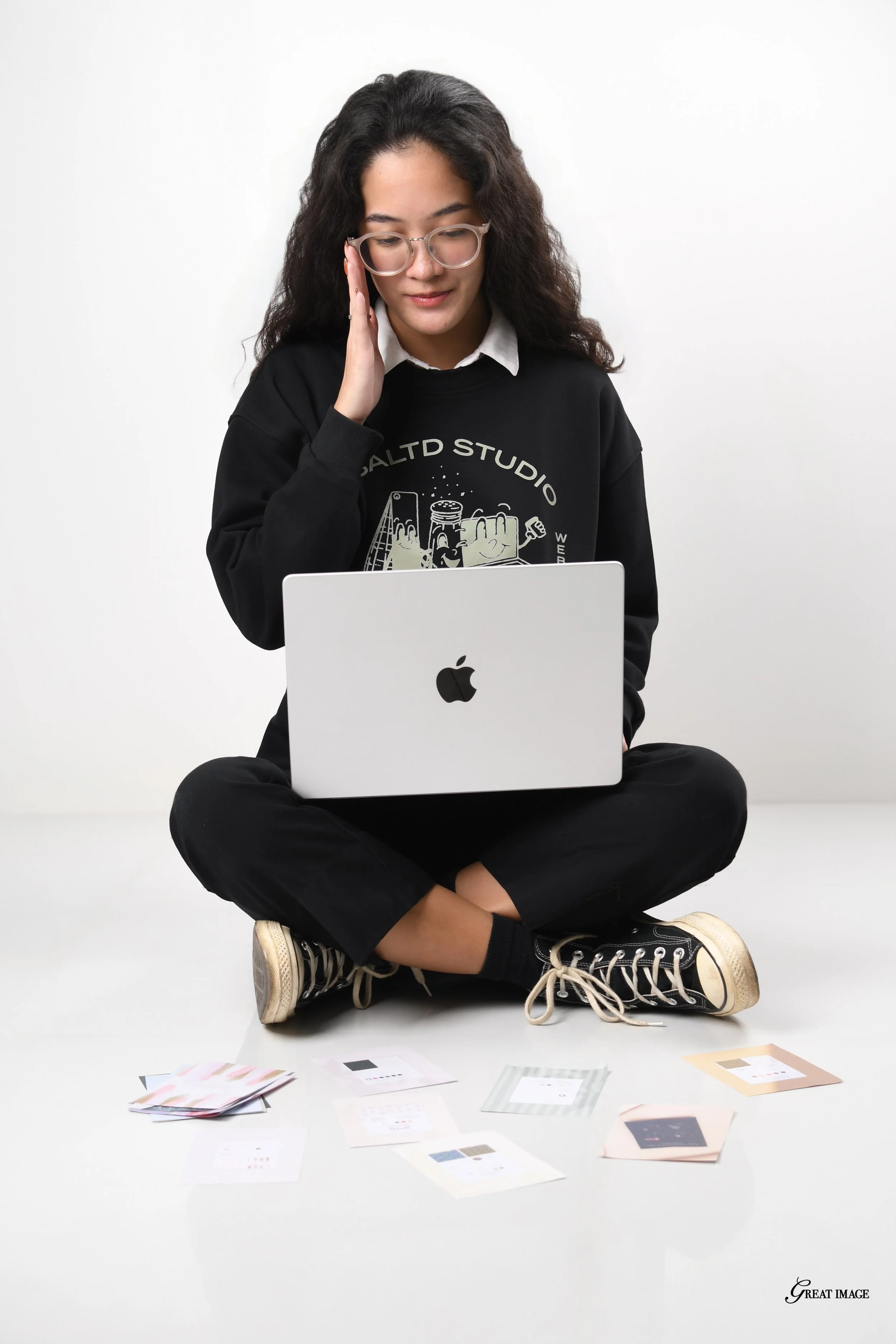 A young woman with curly hair, glasses, and casual clothing, sitting cross-legged on the floor in front of a laptop, with printed paper samples scattered around her. She appears to be analyzing or reviewing printed material.