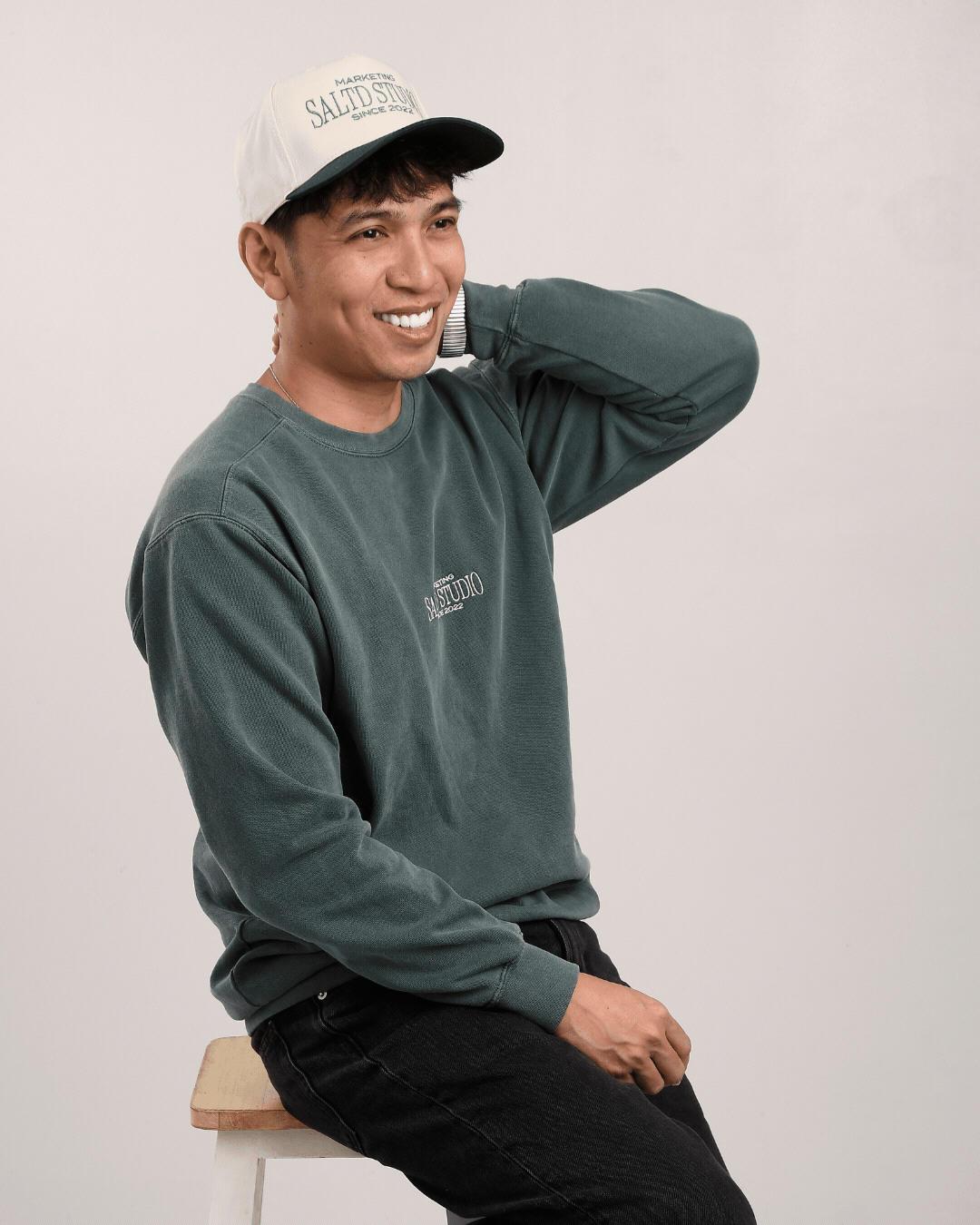 A smiling man wearing a green sweatshirt and a white and black cap, sitting on a stool against a plain background.