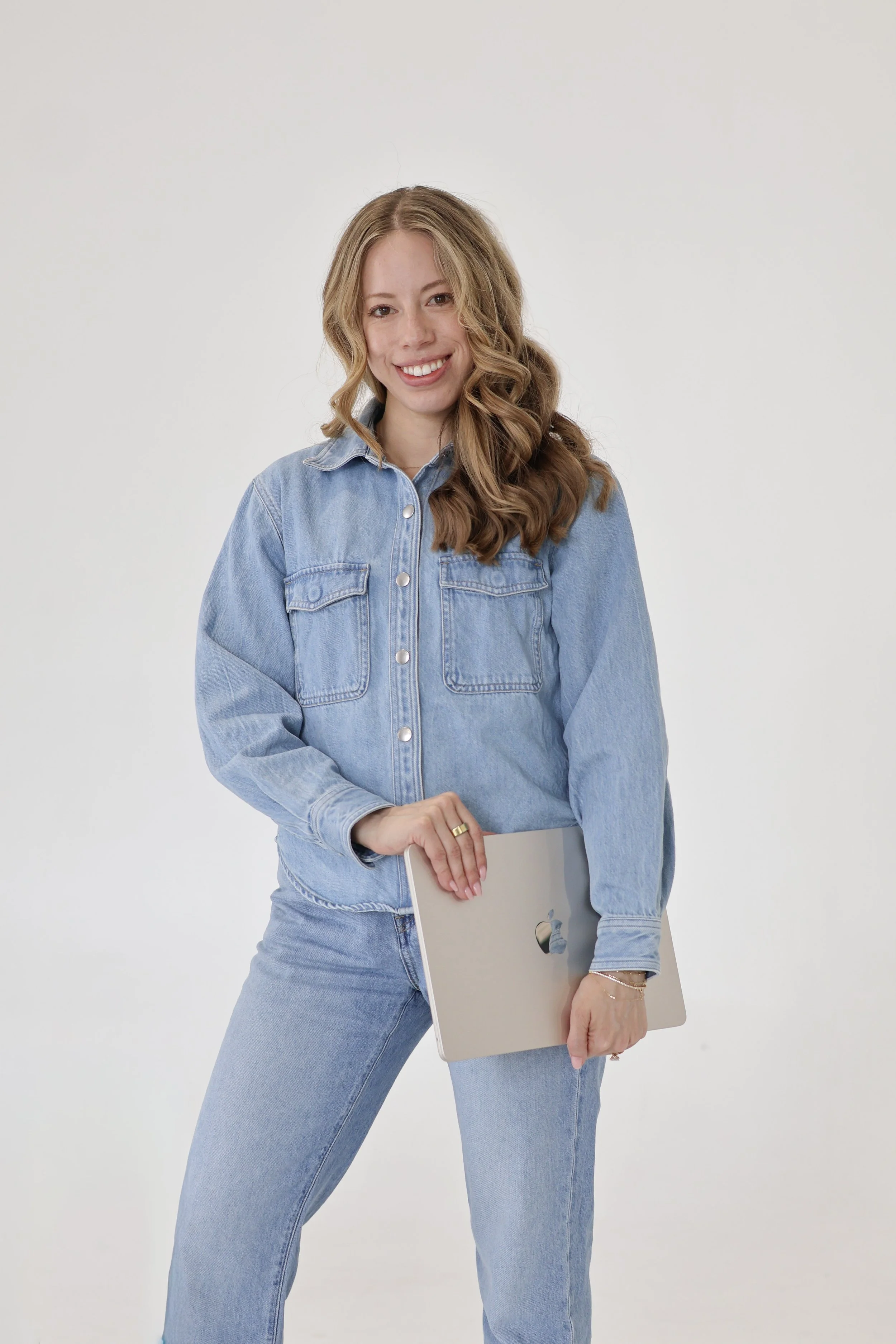 A woman with wavy blonde hair wearing a light blue denim jacket and jeans, holding a silver MacBook laptop, standing against a plain white background, smiling.