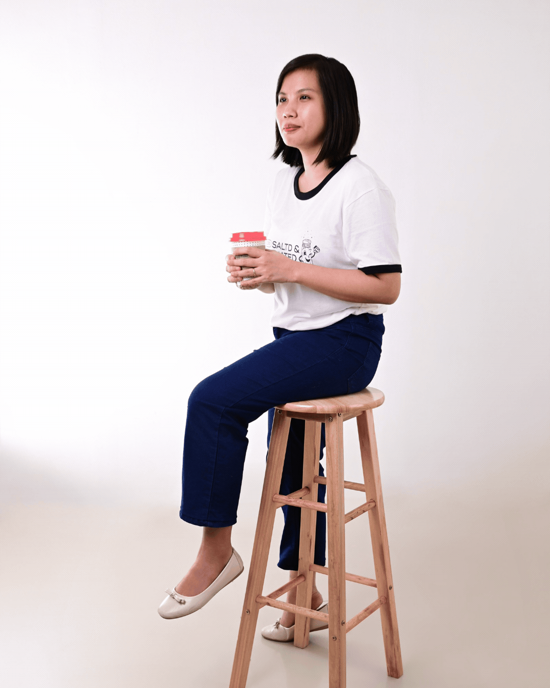 A woman sitting on a wooden stool, holding a mug with both hands, wearing a white t-shirt and dark blue jeans, with a neutral background.