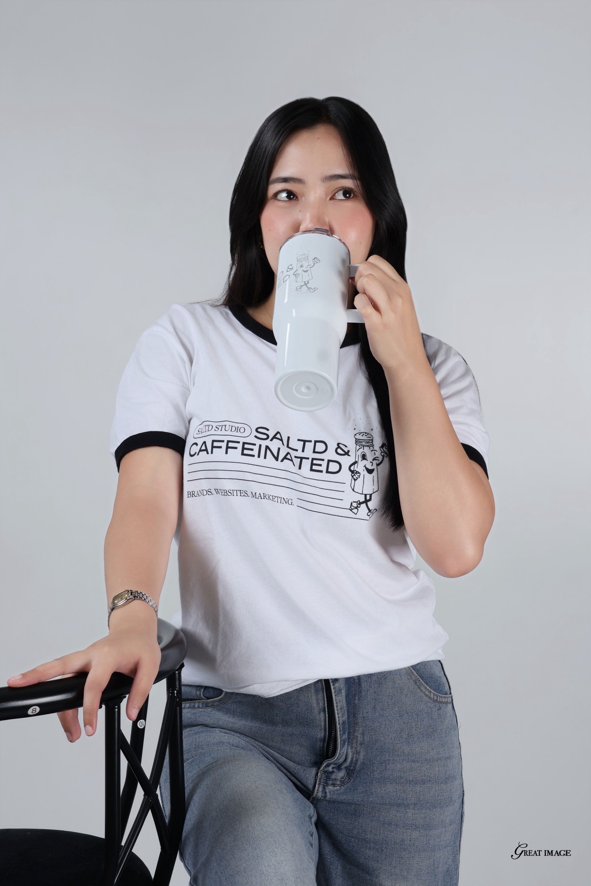 A woman with black hair wearing a white t-shirt is drinking from a white mug with a cartoon character on it. She is also holding the handle of a wheelchair. The background is plain gray.